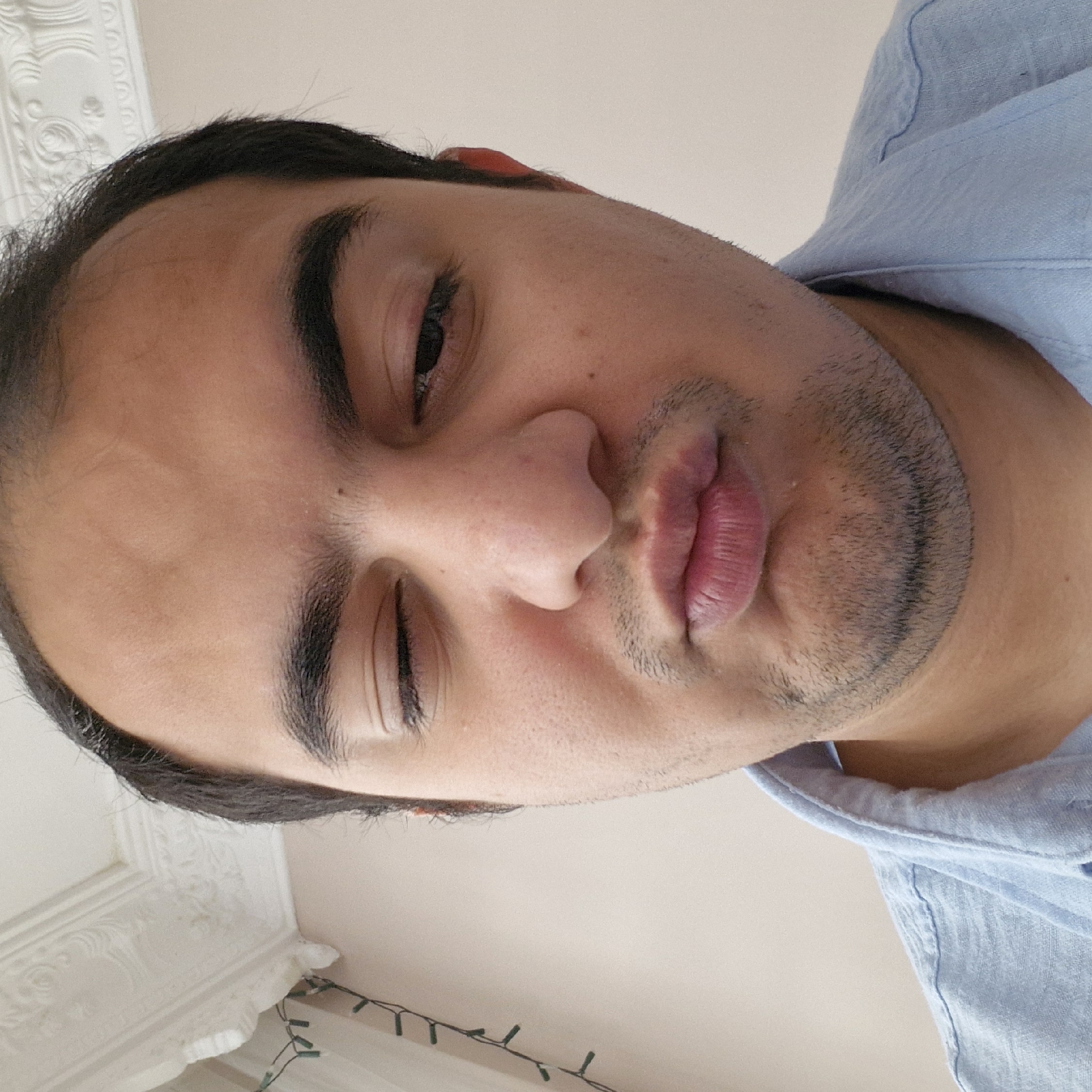 The image is a close-up, selfie-style photograph of a person's face, taken indoors against a light-colored wall with decorative molding at the top. The subject is winking with their left eye and pursing their lips, suggesting a playful or flirtatious expression. The person is wearing a light blue collared shirt. The background shows a string of small, green-lit fairy lights draped along the wall to the left of the frame, and ornate white molding along the top edge. The lighting is natural and appears to be coming from the front, casting soft shadows on the face. The image is cropped tightly, focusing on the upper portion of the face, including the eyes, nose, and mouth, with the shirt collar visible at the bottom. There is no discernible indication of the specific location within Küçükçekmece, Turkey.