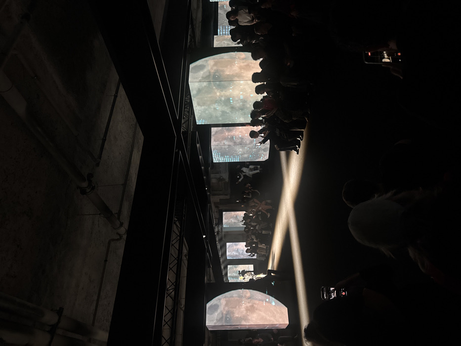 The image depicts an indoor event, likely a fashion show or presentation, held in a large, dimly lit industrial-style space within the **Palais de Tokyo** complex, located in the 16th arrondissement of Paris, France. The environment features a raw, exposed concrete ceiling with visible industrial pipes and dark, heavy structural beams. Multiple vertical rectangular screens are positioned along both sides of the event space, displaying dynamic imagery that appears to be a celestial body (like a moon or planet) overlaid with abstract or urban patterns.
In the center of the scene, a well-defined rectangular spotlight illuminates a dark-colored floor, forming a runway. A single individual, possibly a model, is visible in the distance at the end of this illuminated path, facing away from the camera. Numerous attendees are seated in rows on either side of the central area, mostly silhouetted against the ambient light from the projections and the spotlight. Many individuals in the audience are actively holding up smartphones, suggesting they are recording or photographing the ongoing event. The overall atmosphere is dark and dramatic, with focused lighting on the central presentation area and immersive visual projections.