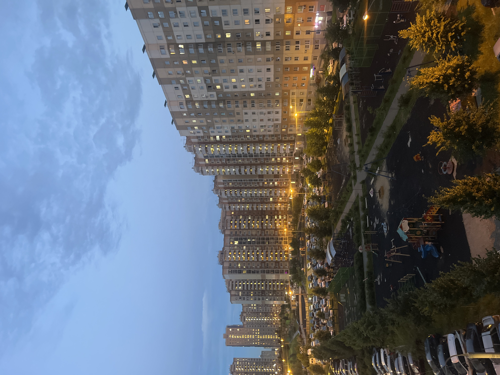 The image depicts a view of a residential area in Domodedovo, Russia, during twilight. Dominating the scene are multiple tall, multi-story apartment buildings, their windows illuminated, indicating occupancy. In the foreground, a children's playground is visible, featuring various play structures such as swings, slides, and climbing equipment, with a sandpit in the background. Adjacent to the playground is a parking lot filled with numerous cars, suggesting a densely populated urban environment. Trees and green spaces are interspersed throughout the area, providing a contrast to the concrete structures. The sky is overcast with a gradient of blues and grays, typical of the transition from day to night. The lighting suggests that artificial streetlights are also present, casting a warm glow on some of the pathways and play areas. There are no discernible individuals in the image engaged in any specific actions.