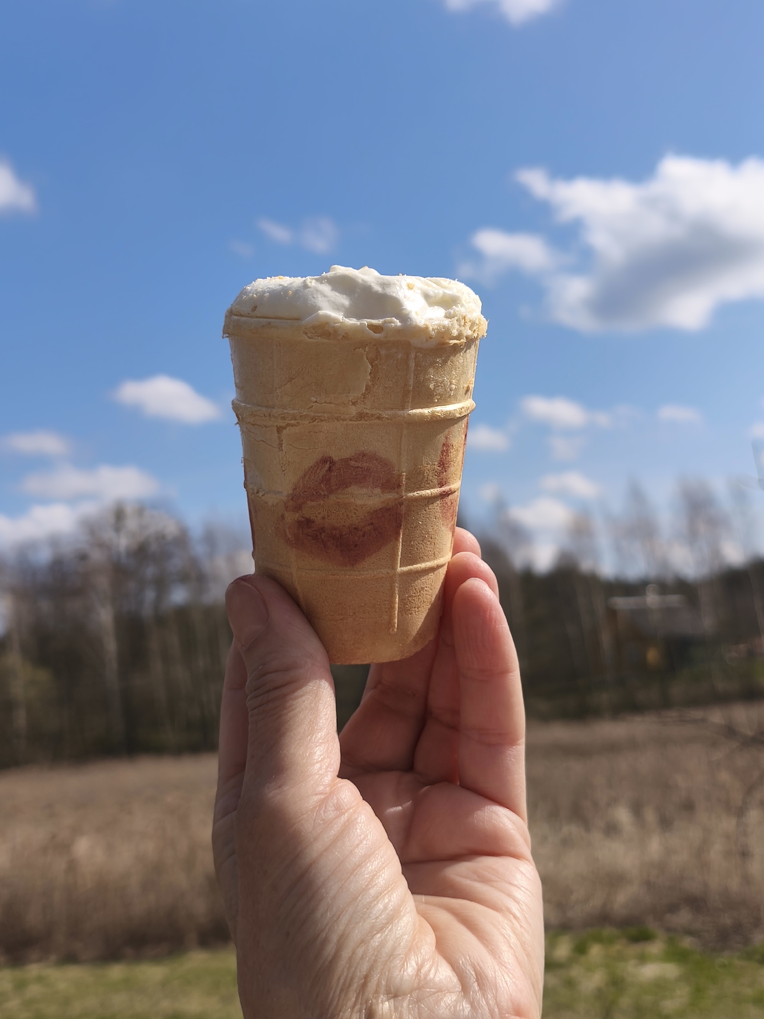 A hand holds an ice cream cone with a lipstick mark on it. The background shows a field, trees, and a blue sky with clouds. The location is Парэчча, Belarus.