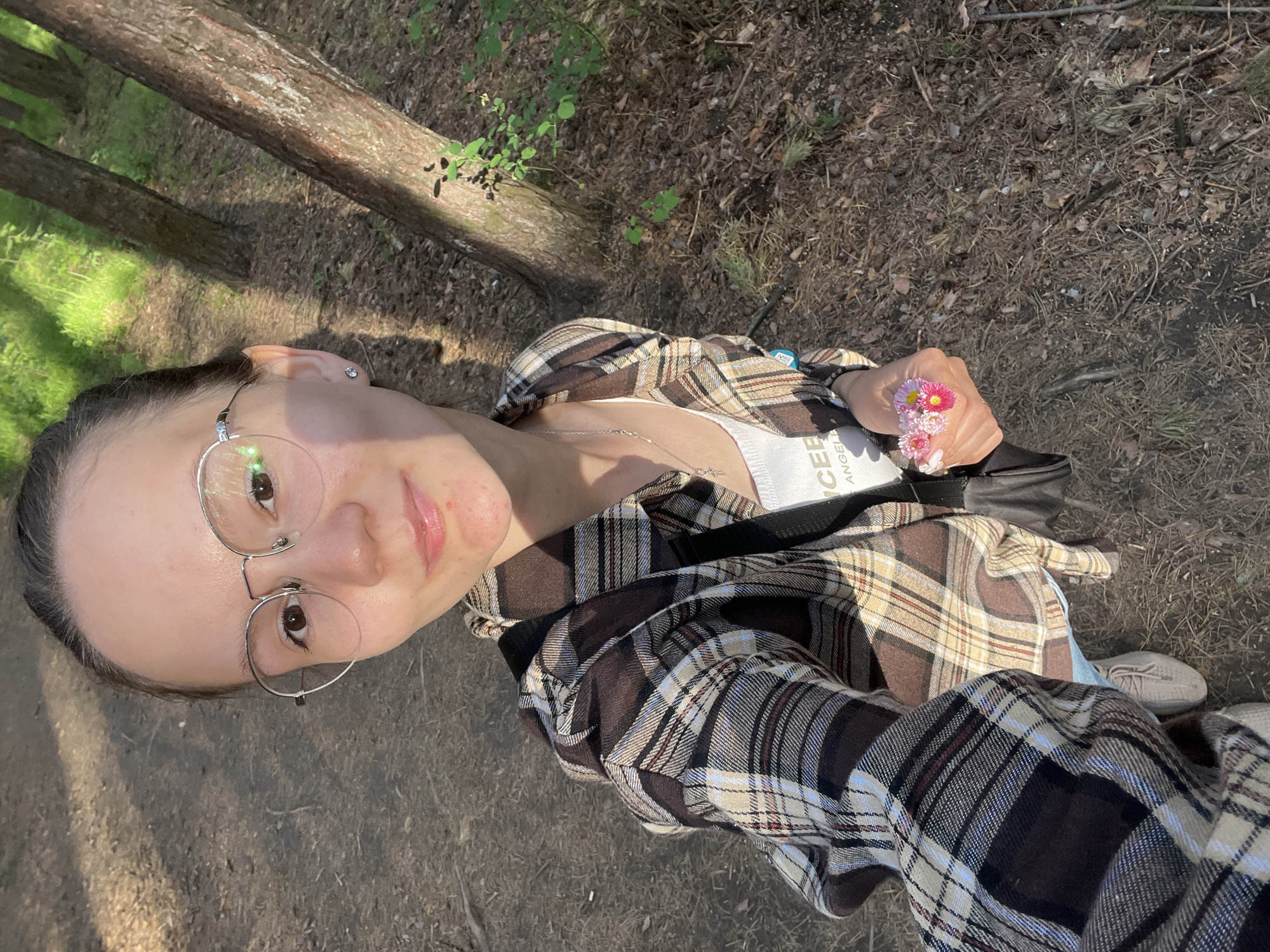 A selfie taken by a young woman with light skin, wearing silver wire-rimmed glasses, a white tank top, and a brown plaid flannel shirt. She is holding a small bouquet of pink and white flowers in her right hand. A black purse is slung across her body. The background is a forest floor with fallen leaves and a tree trunk in the upper left corner. A portion of a dirt path is visible on the left. This image appears to be taken outdoors, likely in a park or wooded area near the city of Chaikovsky, Russia.