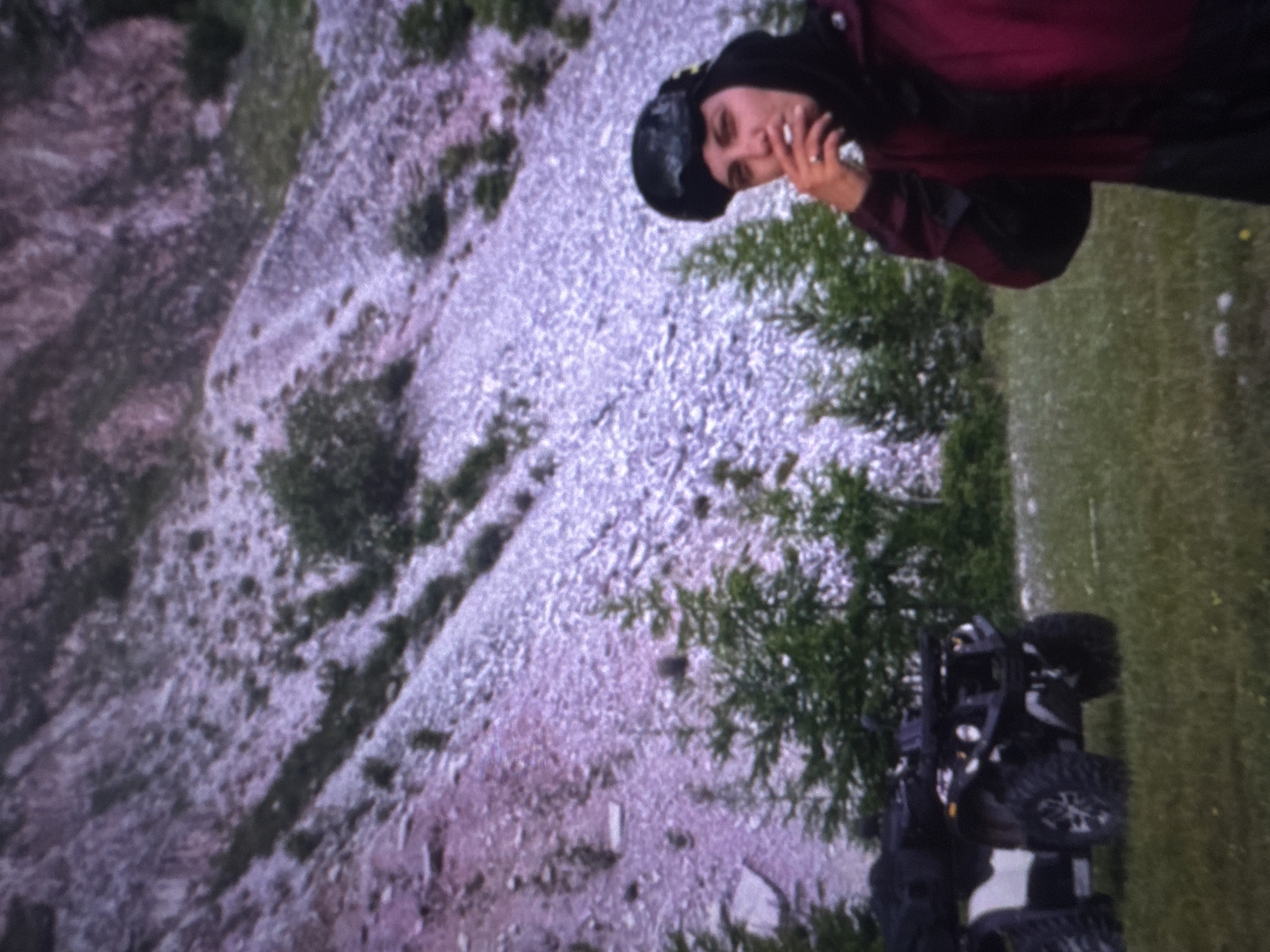 This media file depicts a person in a dark beanie and a burgundy jacket standing next to a black ATV. The person appears to be holding a cigarette to their lips and is looking towards the camera. They are wearing a ring on their left hand. The background consists of a rocky, uneven terrain with scattered green vegetation, and a grassy area to the right. The scene is outdoors, suggesting a rural or wilderness setting. The lighting and general appearance of the image indicate it might be daytime, though the specific time is not discernible. The weather appears to be overcast or possibly rainy, given the muted colors and the person's attire. The presence of the ATV suggests an activity like off-roading or recreational travel in a natural environment. The provided location context of Irkutsk, Russia, places the scene within a specific geographical region known for its varied landscapes. There is no visible text in the media file.