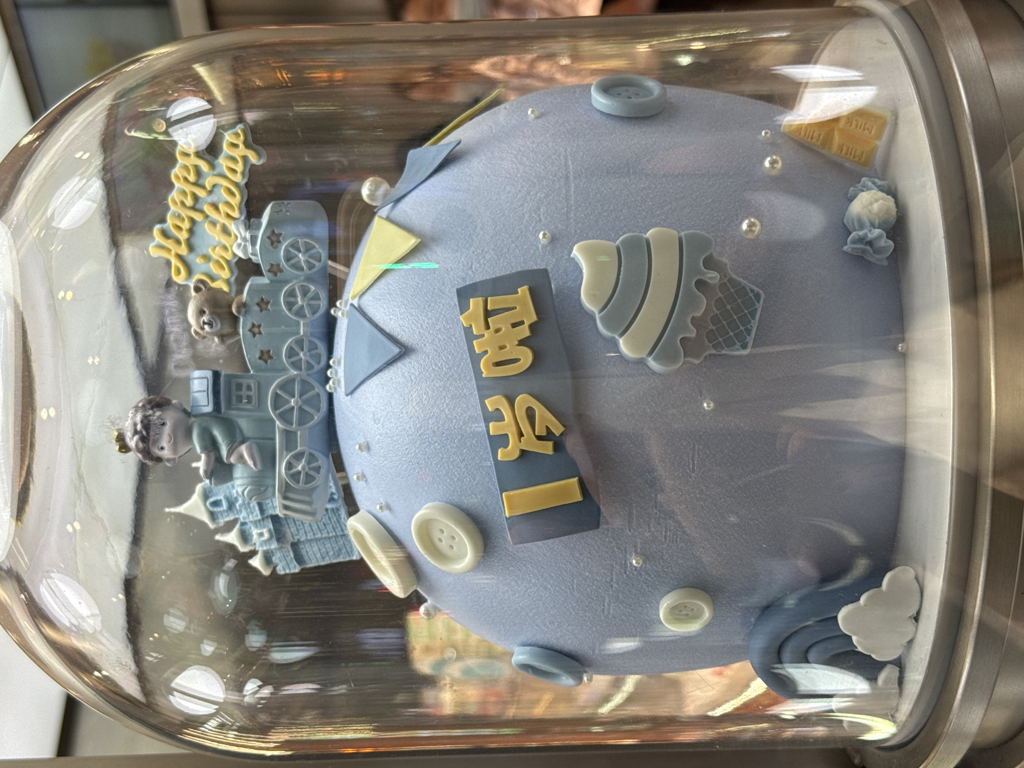 A blue cake with a train decoration is displayed inside a glass dome. The cake is decorated with small buttons and other decorative elements. The scene appears to be indoors. "Happy Birthday" is written on the side. It's located in Qianjiang, China.