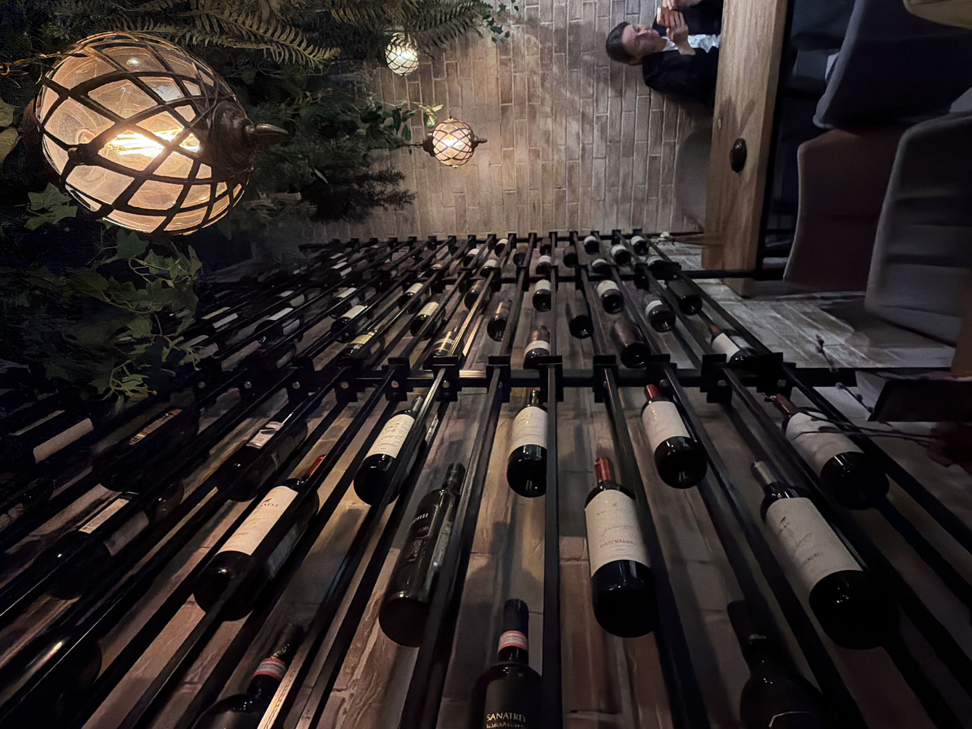 The image depicts an indoor wine rack in what appears to be a restaurant or bar. Several rows of wine bottles are displayed horizontally within a black metal rack system. A lit, caged light fixture hangs on the left side of the frame, near some greenery. In the upper right corner, a man is seen holding a phone, possibly taking a picture. A partially visible wooden doorframe and some chairs are also in the background. The lighting is dim, creating a warm, inviting atmosphere. There is no information to deduce if the location is in Izhevsk, Russia.
