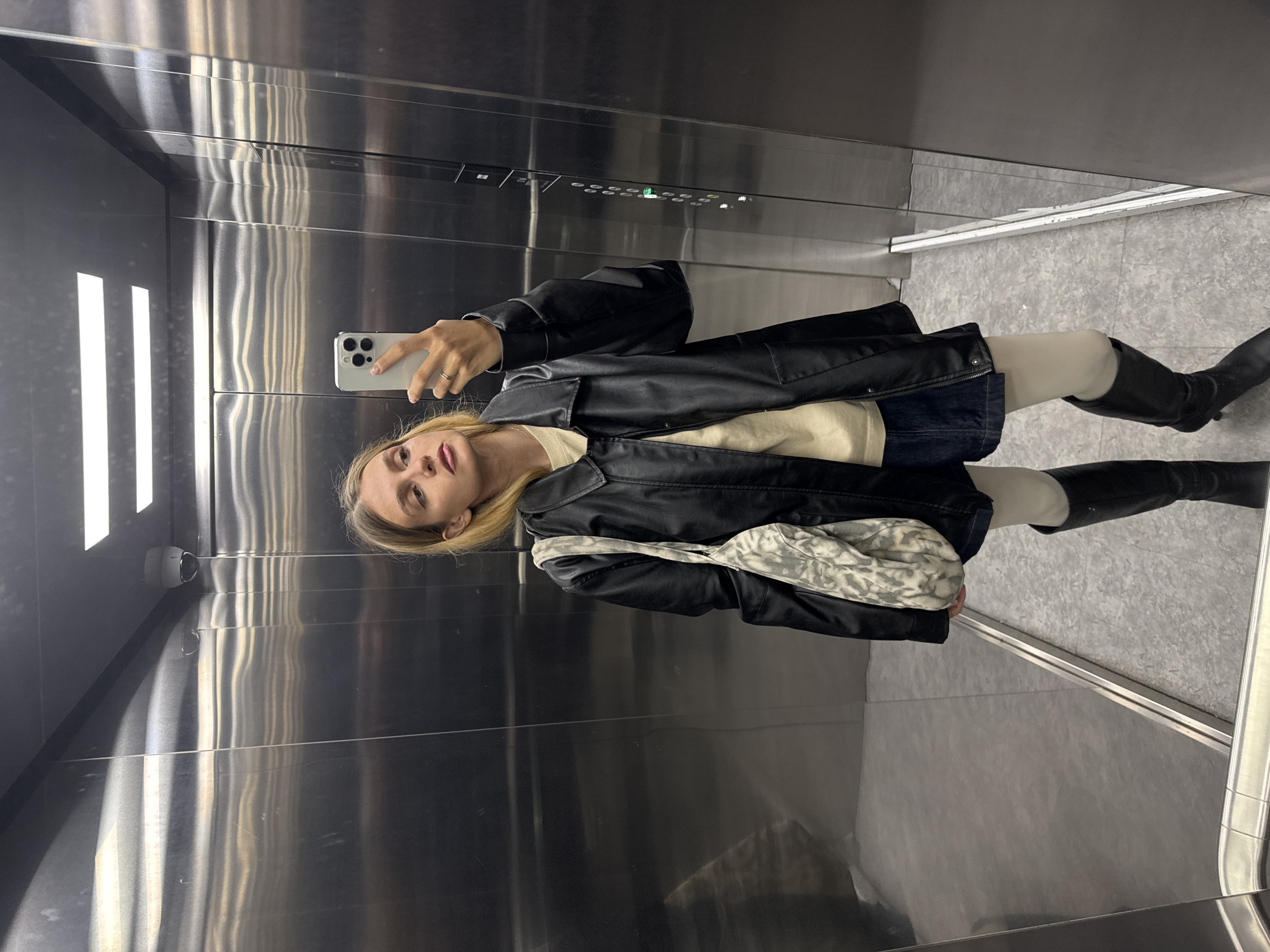 A fair-skinned person with long blonde hair stands in a stainless steel elevator, taking a selfie with a white smartphone. The person is wearing a black leather jacket, a beige sweater, a dark denim skirt, pale tights, and black knee-high boots with heels. A patterned fabric bag hangs from their shoulder. The elevator's interior includes a panel with buttons, a security camera, and artificial lighting. The floor is gray with a metallic edge. The scene appears to be a reflection in the elevator's mirrored wall. The location and city are unknown.