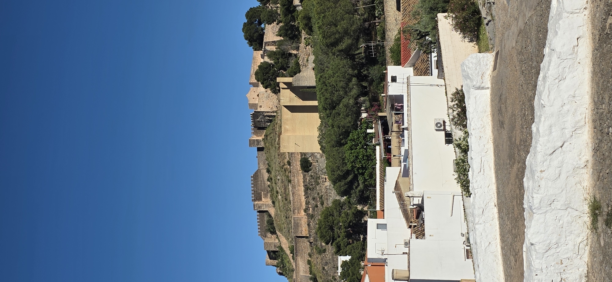 The image depicts a landscape scene with a historic castle in Sagunto, Spain, positioned on a hilltop. The sky is clear and blue. The castle is built with stone walls and defensive structures and partially covered by green trees and vegetation. Adjacent to the castle hill, there are white buildings with tiled roofs and balconies, situated along a white-painted stone road, suggesting a residential area. An air conditioning unit is visible on the wall of one building. The scene includes a mix of ancient architecture and modern dwellings.