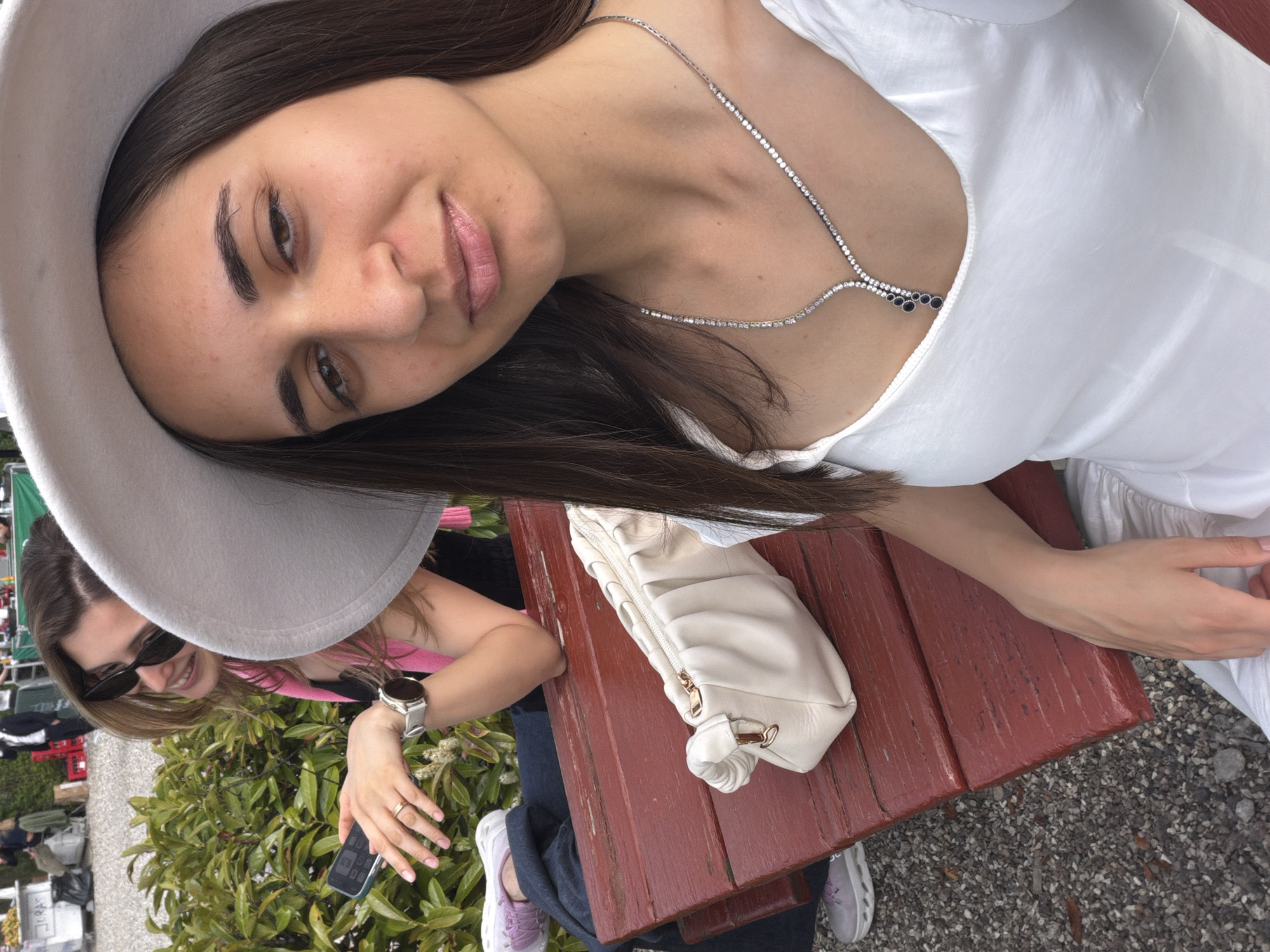 The image shows a woman wearing a wide-brimmed hat and a white dress sitting at a wooden table. She has a silver necklace. Next to her on the table is a white purse. In the background, another woman with sunglasses is visible next to a leafy bush. The location appears to be outdoors.