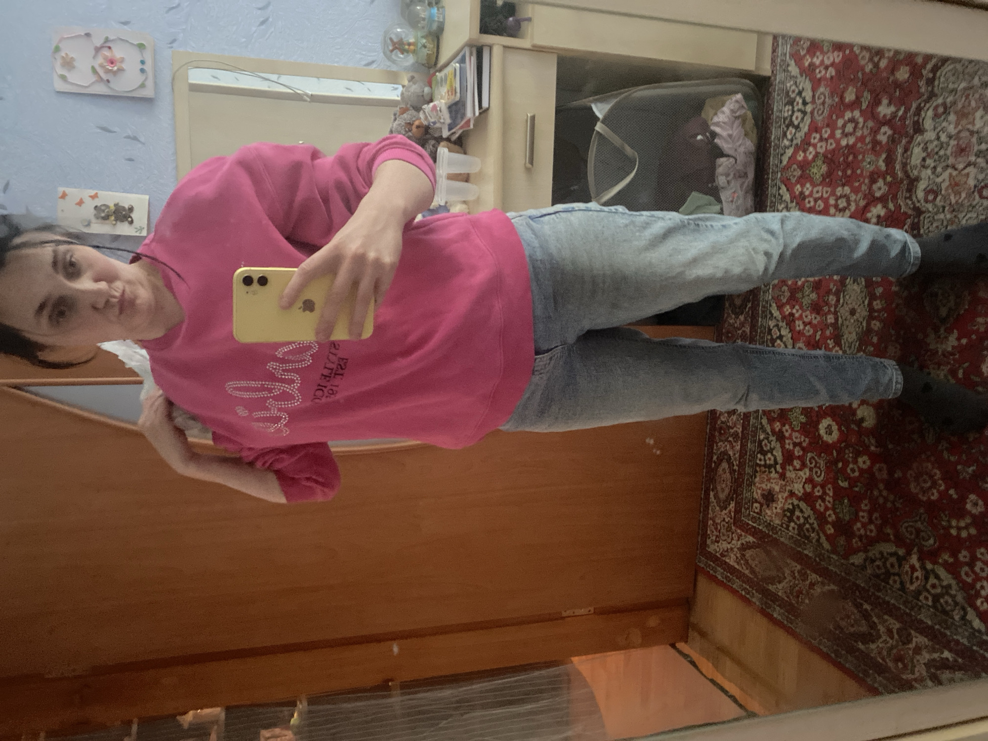 A full-body mirror selfie shows a woman with dark hair, pulled back loosely, wearing a bright pink long-sleeved sweatshirt with a bejeweled graphic and light-wash denim jeans. She is holding a yellow smartphone in her right hand, capturing the image. The background reveals a bedroom setting with a wooden door on the left, a vanity table with a mirror on the right, decorated with various items including small figurines and books. A patterned rug covers the floor. The lighting appears to be indoor, likely from artificial sources. The overall scene is casual and personal, suggesting a domestic environment.
