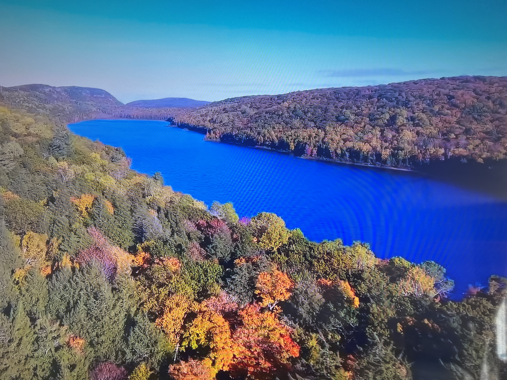 The image shows an aerial view of a long, narrow body of water, possibly a lake or reservoir, surrounded by forested hills. The water is a deep blue color. The hills are covered in trees displaying a mix of autumnal colors, including green, yellow, orange, and red. There are mountains in the background. The sky is a clear light blue. There are no people visible in the image. The location is unspecified, but given the fall foliage and topography, it is likely a temperate region. It is not known to be Basingstoke and Deane, United Kingdom.