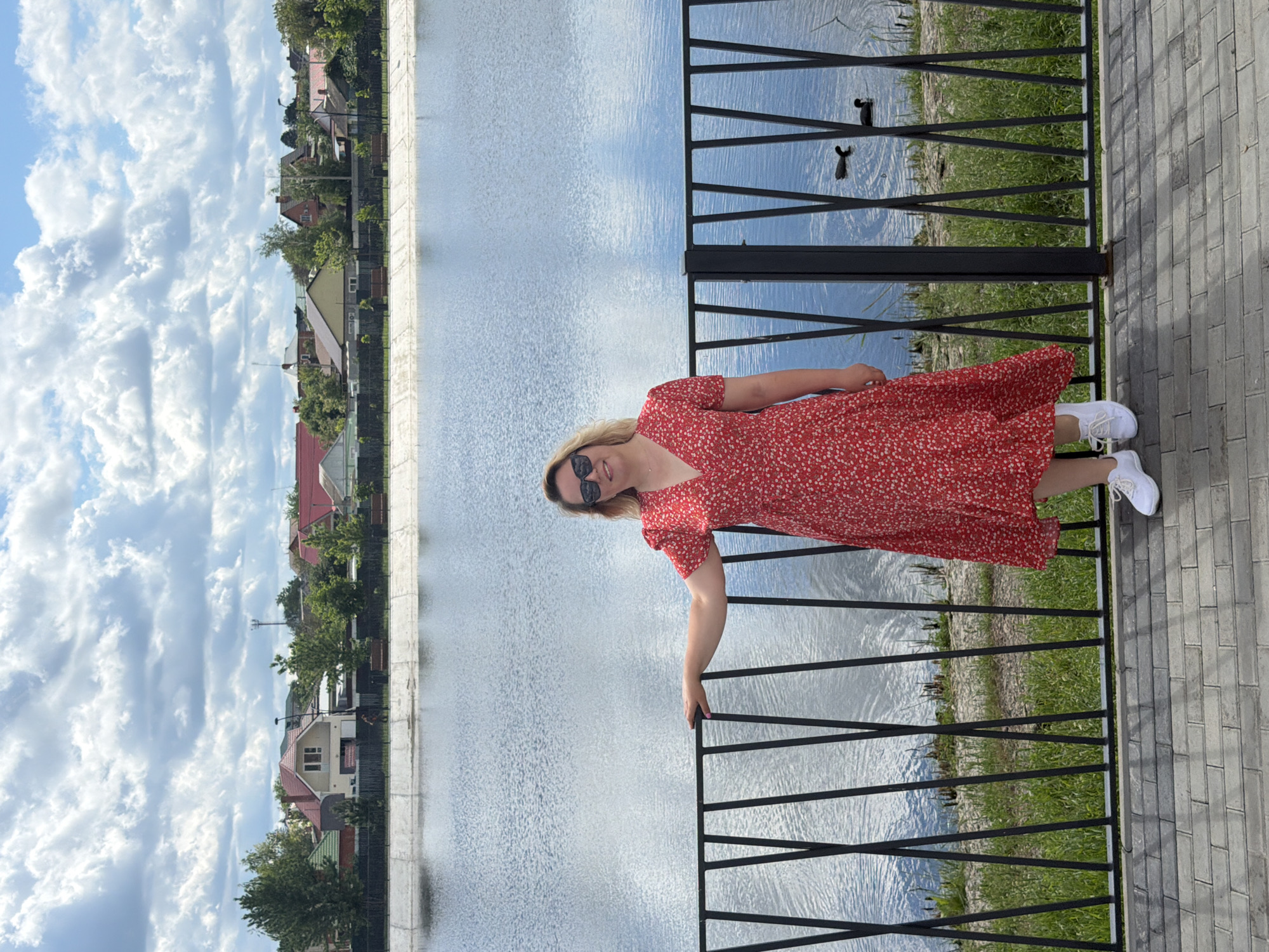 A woman is standing on a brick walkway beside a black metal railing that borders a body of water. She is wearing a red dress with a small floral pattern, sunglasses, and white sneakers. The water reflects the sky and some of the surrounding buildings. On the opposite side of the water, there are buildings, some with red roofs, and trees. Above, there is a blue sky with fluffy white clouds. The scene is likely in the city of Leningradskaya, Russia, as indicated in the prompt.