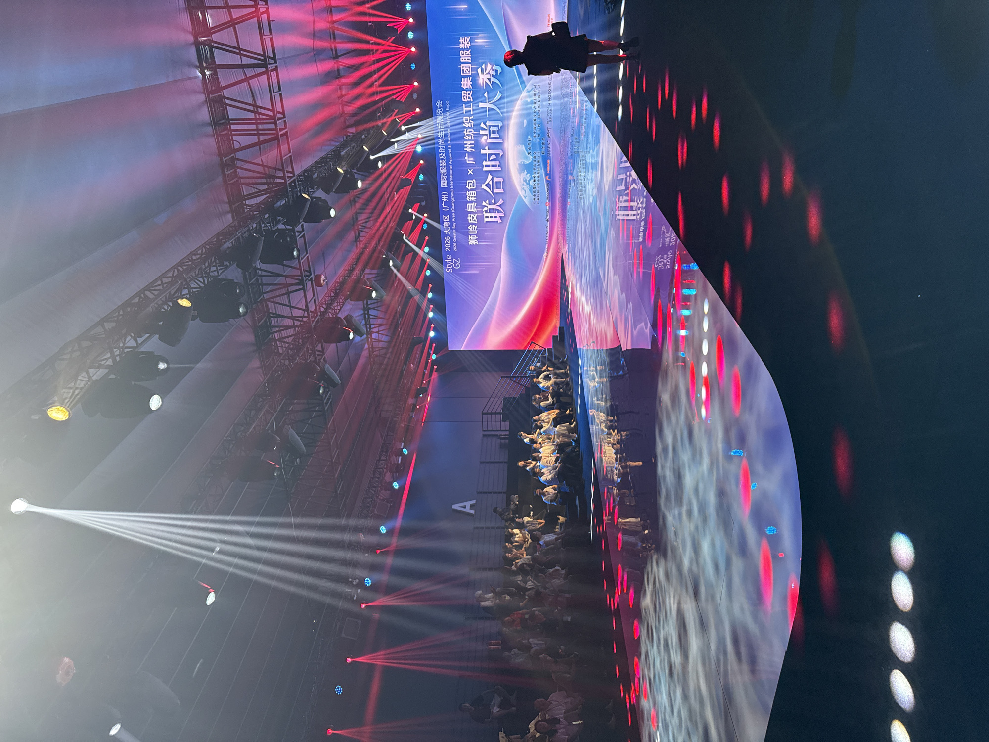 The image captures a fashion show event taking place at an indoor venue, likely in Haizhu District, China, given the context of the "Style 2026 Greater Bay Area (Guangzhou) International Apparel & Fashion Lifestyle Expo." The scene is dominated by dramatic stage lighting, with bright white and vibrant red beams cutting through the smoky air. A large screen displays colorful graphics and text, including the event title in Chinese characters: "联合时尚大秀" (United Fashion Grand Show). In the foreground, a dark, reflective stage floor is illuminated by red and pink circular light patterns, creating a dynamic visual effect. On the right side of the stage, a lone figure, possibly a participant or staff member, stands facing the screen, dressed in black with a red head covering. To the left, an audience of people is seated on tiered risers, observing the show. The overall atmosphere is one of high energy and visual spectacle, typical of a large-scale fashion event.