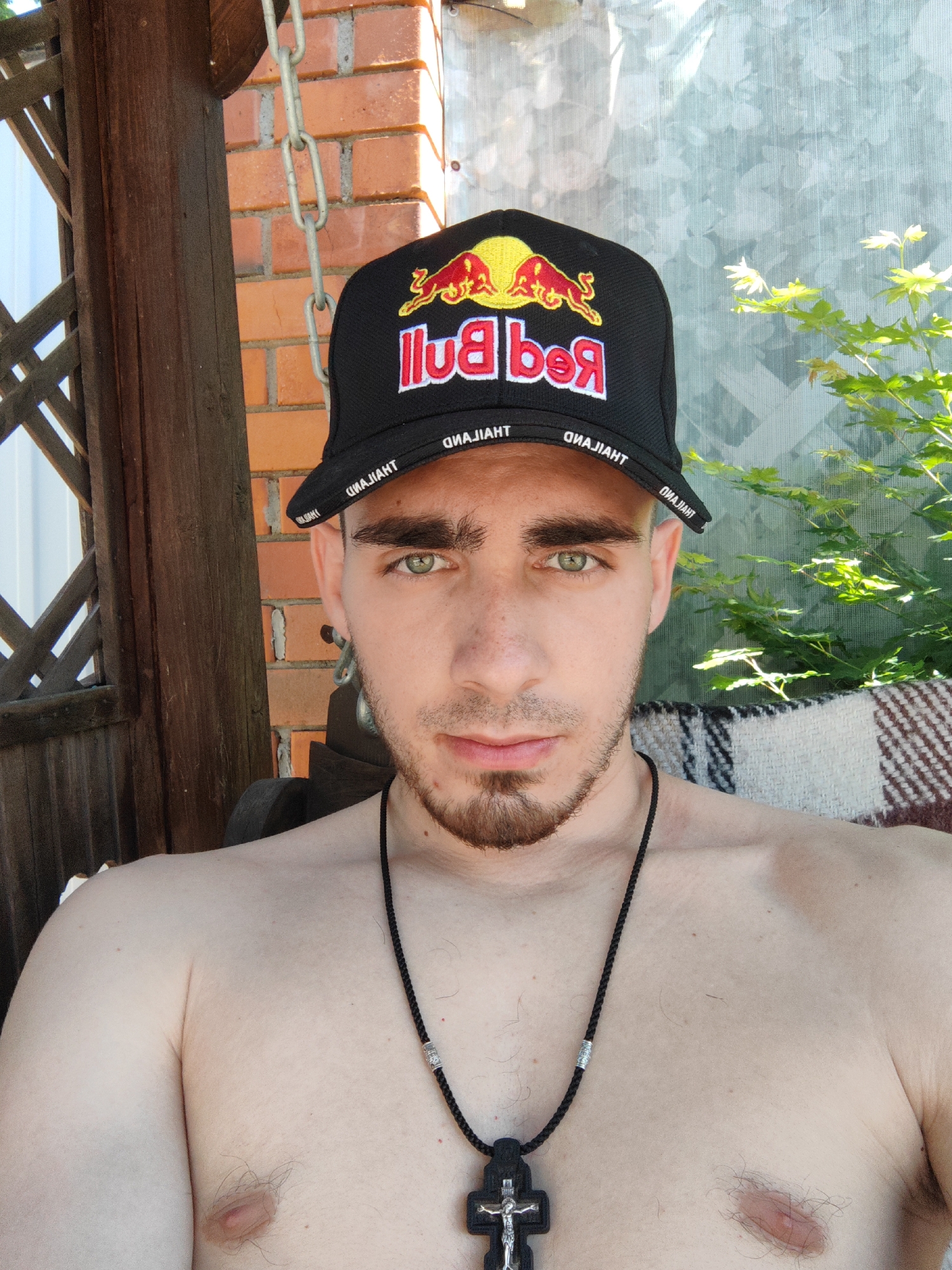 A selfie shows a young man, appearing to be of Caucasian ethnicity, with light skin, green eyes, dark brown hair, and a short beard. He is wearing a black baseball cap with the Red Bull logo and "THAILAND" written on the brim. He is also wearing a black necklace with a silver bead and a large, black, crucifix pendant. The man is shirtless, revealing his chest and shoulders. He appears to be sitting outside on a patterned blanket. Behind him, there is a brick wall, a wooden structure, and some green foliage. The scene suggests a casual, outdoor setting, possibly a backyard or patio. There is no specific information available about the location being in the city of Ussuriysk, Russia.