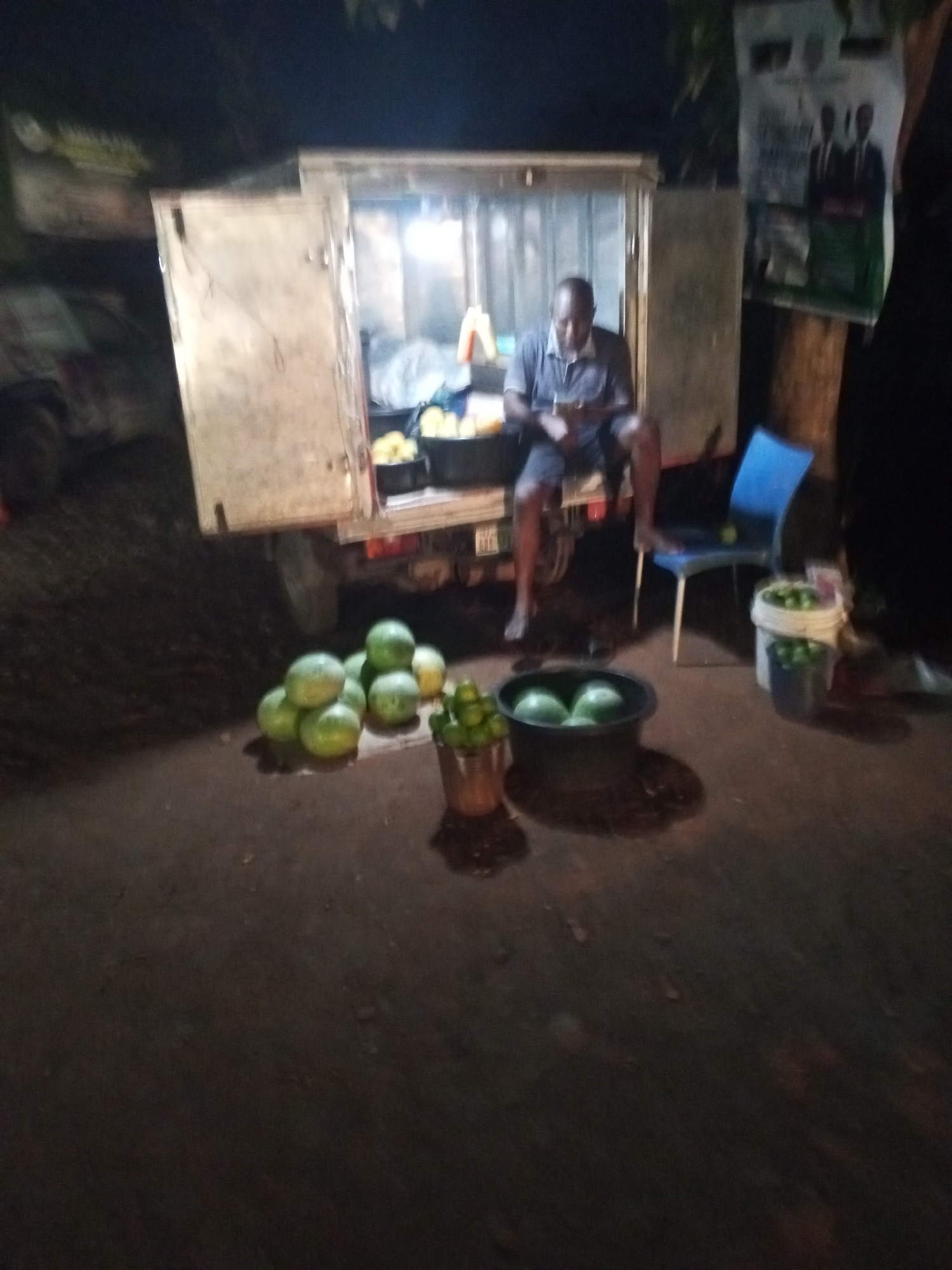 In Galadimawa Village, Nigeria, a man sits on the back of a parked truck with the doors open, lit from inside. He is barefoot and wearing a short-sleeved shirt and shorts. Buckets and piles of green fruits, possibly watermelons or similar produce, sit on the ground nearby. A blue plastic chair is to the right of the man, and a poster is visible on a pole behind the chair. The scene appears to be at night.