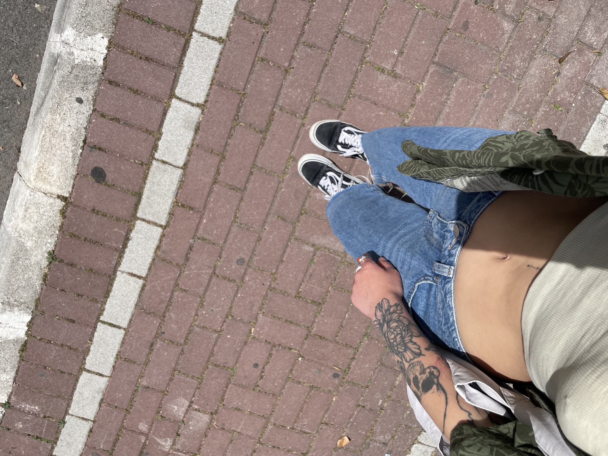 A person wearing blue jeans and black and white sneakers is standing on a brick walkway with a white concrete curb to the left. The person is wearing a white shirt and a green patterned shirt. They have tattoos on their left arm. The scene appears to be outside in daylight. No evidence indicates the location of Rafailovići, Montenegro.