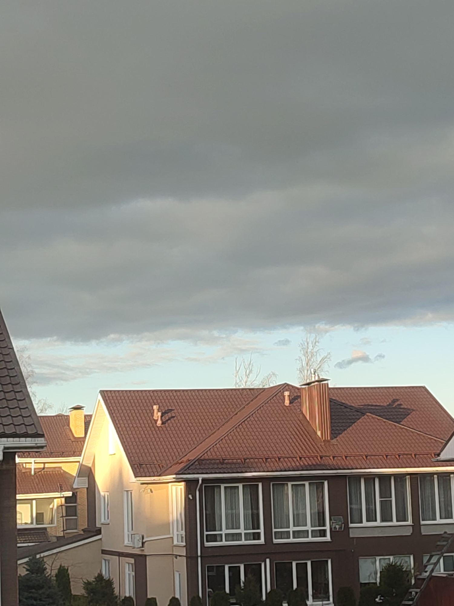 This image presents a residential scene in Zagoryanskiy, Russia, depicting several modern houses under a partially cloudy sky. The main subjects are the multi-story houses, characterized by their prominent brown tiled roofs, varying facades, and numerous windows.
The sky occupies a significant portion of the upper frame. It is largely covered by a thick, grey layer of clouds, suggesting overcast conditions. However, a band of lighter sky, with some wispy clouds and hints of blue, is visible lower down, just above the rooftops, indicating a break in the cloud cover or clearing weather.
The houses exhibit distinct architectural features. Most have complex brown tiled rooflines with multiple gables and visible chimneys; one notable chimney appears to be constructed from a metallic material. The house facades vary, with some displaying light beige or pale yellow rendering, while others feature darker brown sections, particularly on their lower levels. Numerous large windows with white frames are present across the buildings. One of the lighter-colored houses is conspicuously illuminated by golden sunlight on its side, casting a sharp shadow line, which suggests the time of day is either early morning or late afternoon. Leafless tree branches are visible against the sky and between the houses, hinting at a cooler season. Evergreen bushes and shrubs are planted around the base of the houses.
No human activity or individuals are visible in the tranquil scene. There is no discernible text on any of the visible objects or buildings.