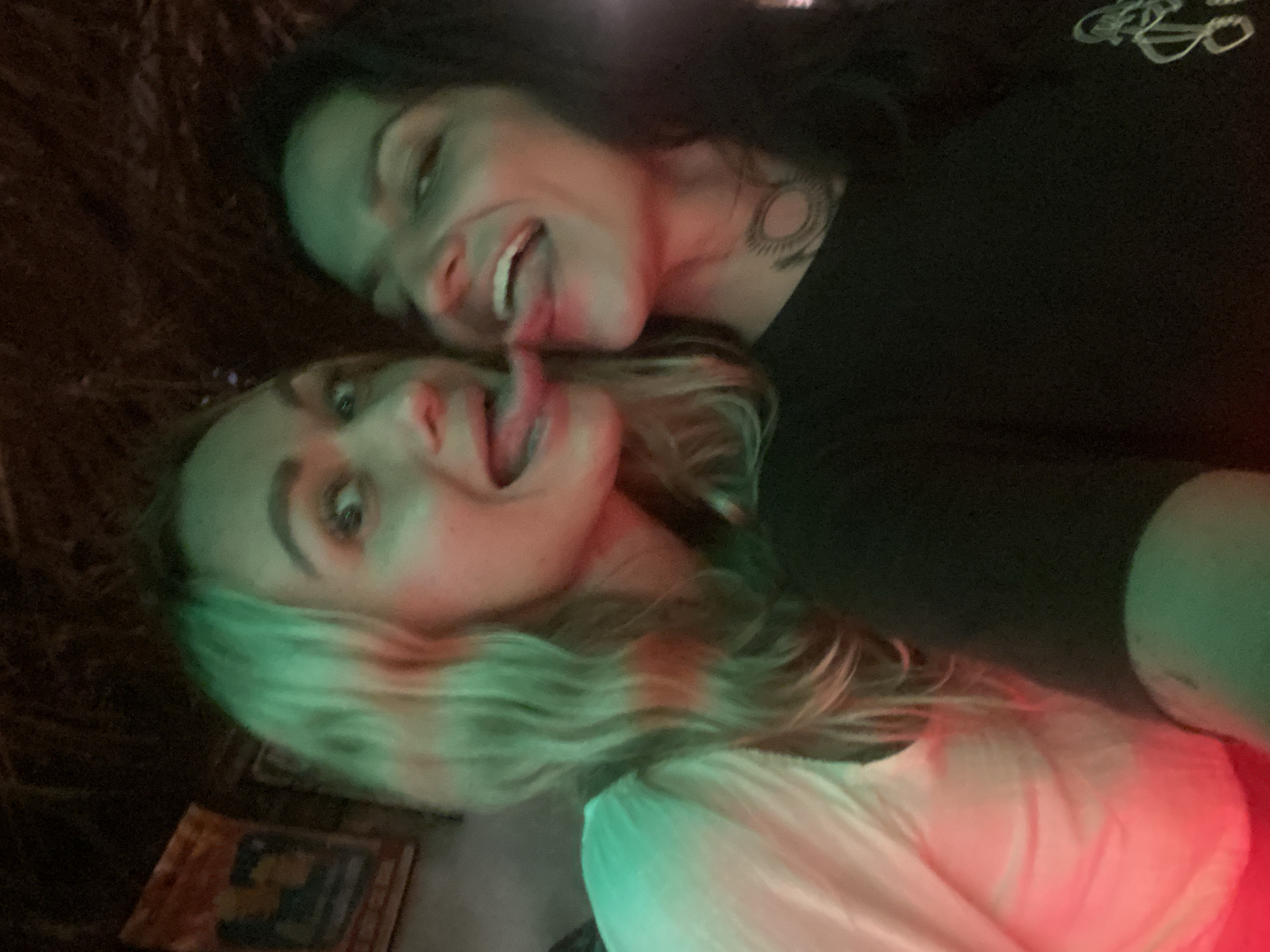 This media file captures a rotated, close-up photograph of two individuals, likely women, in what appears to be a dimly lit indoor setting, possibly a club or bar in Mũi Né, Vietnam. The image suggests a night-time setting due to the prominent use of artificial, colored lighting.
The two subjects are positioned closely, engaging in a playful moment for the camera. The person positioned higher in the frame has dark hair and is wearing a dark top, with a circular tattoo visible on their neck/shoulder. They are shown with a wide smile and their tongue playfully extended. The person positioned lower has wavy, lighter, possibly blonde hair, and is wearing a light-colored top. They also have a wide smile and their tongue sticking out, mirroring the playful expression of the other individual.
Their faces and hair are illuminated by strong green and red lights, creating a vibrant and dynamic effect on their features, while the background remains dark and textured. The overall mood conveyed is cheerful, informal, and silly. A faint, light-colored, abstract logo or design is visible on the upper right side of the dark-haired person's top. In the lower left part of the image, below the blonde person's head, a small, rectangular object with indecipherable markings is partially visible.