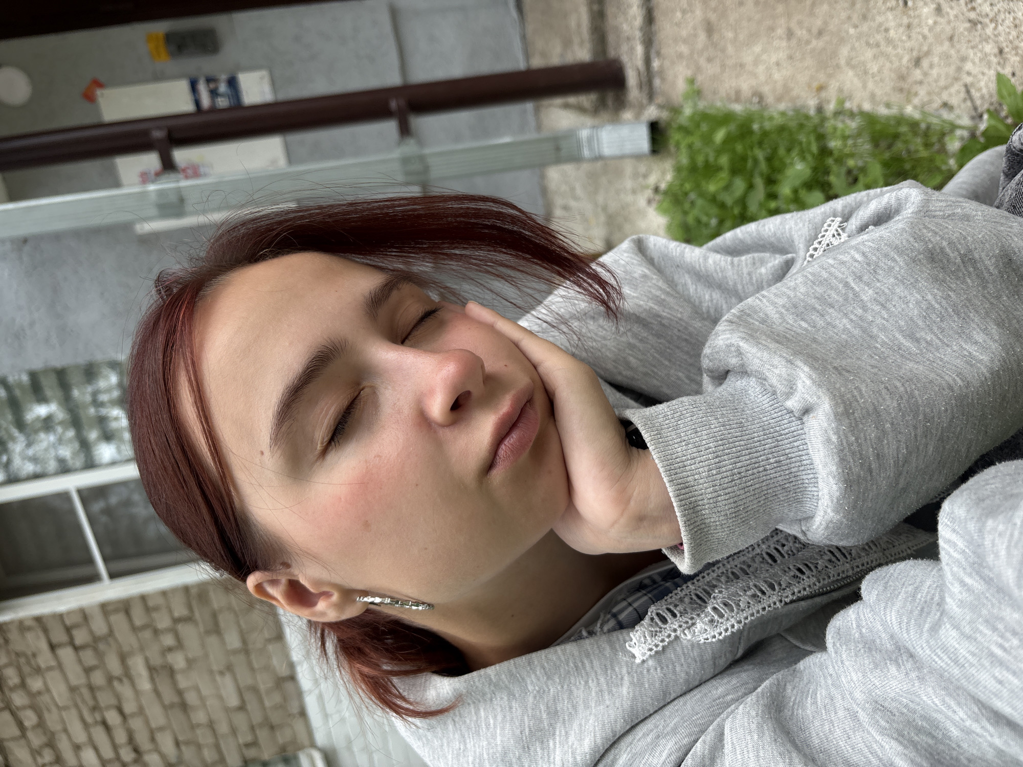 The image is a close-up, selfie-style photograph of a young woman with reddish-brown hair and closed eyes, resting her chin on her hand. She is wearing a gray hooded sweatshirt over a plaid shirt, with a silver hoop earring in her visible ear. The background appears to be an outdoor urban setting, with a brick building and a drainpipe visible. The woman's expression suggests she is tired or contemplative. The location is identified as Chaikovsky, Russia.