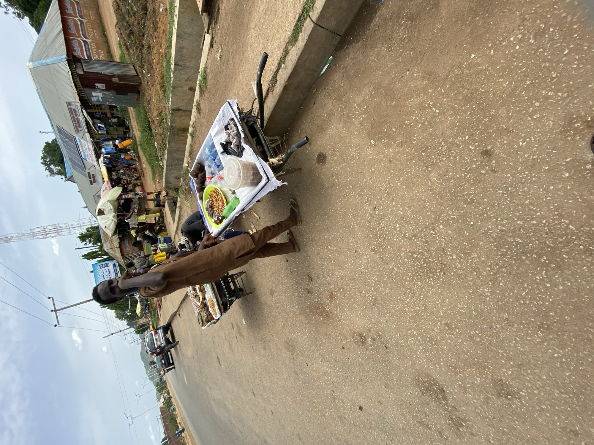 The image depicts a street scene in Bukuru, Nigeria. A man in brown clothing stands near a food cart on the side of a paved road. The cart displays various food items, including snacks and bottled drinks. Nearby, shops and buildings line the street, and a telecommunications tower is visible in the background. The sky is cloudy. Cars are driving in the background.