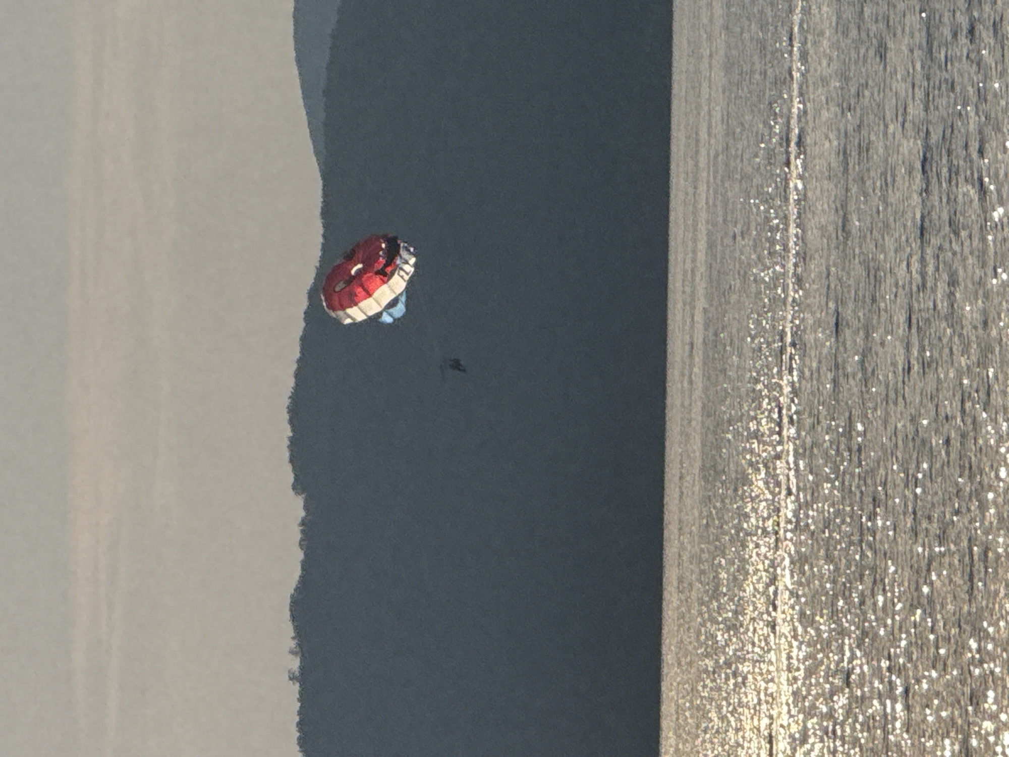 The image shows a parasailer in mid-air, enjoying a recreational activity over a body of water. The parasail is red and white with blue accents, and a person is clearly visible beneath it. The parasailer appears to be enjoying the experience, as evidenced by the upward movement against a backdrop of calm water and a distant landmass. The sun is setting, casting a warm glow on the water, creating a serene and picturesque scene. The water reflects the sky and surrounding landscape, adding depth and beauty to the image. The location appears to be a coastal area, possibly a beach or a bay, with a calm sea and a gentle breeze. The overall mood of the image is one of adventure, freedom, and relaxation, inviting the viewer to imagine themselves in this beautiful setting.