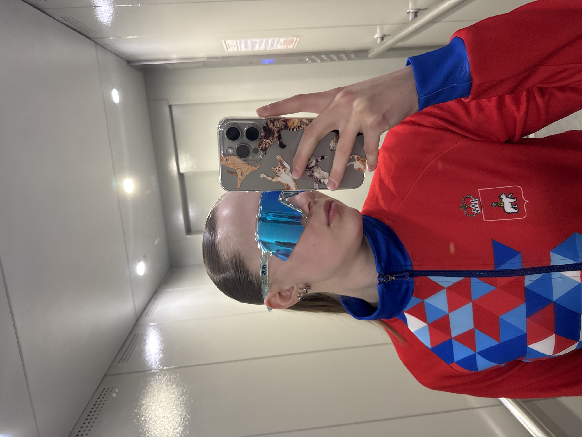 A person is taking a selfie in what appears to be an elevator. The individual is wearing a red jacket with blue accents and a logo depicting a crown and a white animal. They are also wearing blue reflective sports glasses and holding a phone with a cat-themed case. The elevator's interior is white and appears to be well-lit. The location is in the city of Chaikovsky, Russia. The details of the city of Chaikovsky, Russia can be taken from the red jacket.