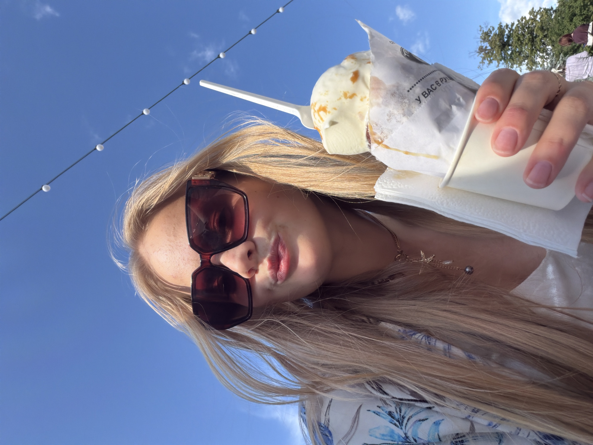 A blonde-haired individual, wearing large dark sunglasses with gold-colored temple accents and a blue and white floral-patterned open shirt over a light undergarment, is centered in the foreground, facing the camera with lips pursed. A delicate gold chain necklace with a starfish charm and a dark pearl-like pendant is visible on their neck. The individual holds a white cup containing a scoop of ice cream topped with caramel sauce and a white plastic spoon, all wrapped in a paper sleeve that displays partially legible Cyrillic text. A white paper napkin is tucked around the cup. The background features a clear blue sky with sparse white clouds, crossed by a string of white spherical lights diagonally from top left to bottom right. In the far right background, green tree foliage and the indistinct figures of two other individuals are visible. The scene depicts an outdoor public setting in the city of Kazan, Russia.