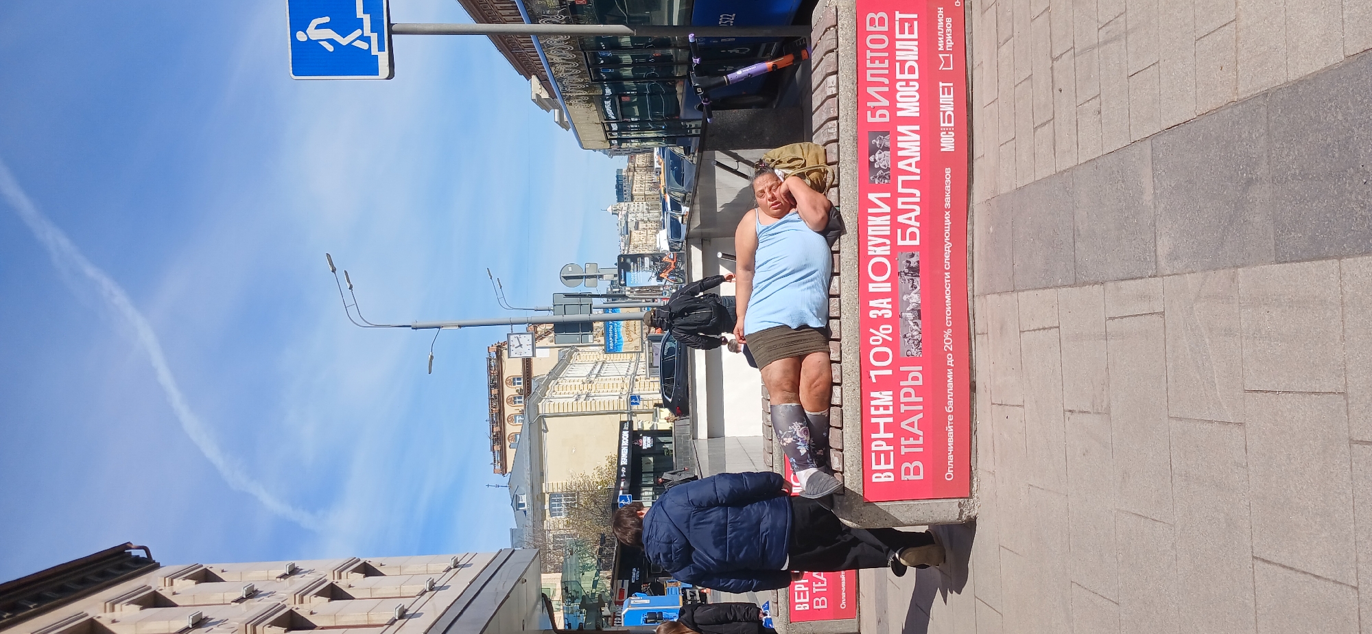 This image captures a vibrant urban street scene, likely in Moscow, Russia, during daytime under a clear blue sky. In the foreground, a person in a dark blue puffer jacket and dark trousers walks away from the viewer on a paved sidewalk. To their right, a large red and white advertising sign stands prominently, with Russian text.
Behind the walking person, a woman in a light blue tank top and dark skirt sits on a low brick wall or bench, her right arm resting on her head, giving a contemplative or weary impression. Her legs are covered with patterned leggings or boots. Adjacent to her is a blue and white public bus (numbered 1322) and two electric scooters.
The background reveals a bustling city environment with multi-story buildings, some featuring ornate architecture and a visible clock face. Streetlights with multiple luminaires extend overhead. Further in the distance, more buildings and vehicles suggest a busy street. A blue pedestrian sign depicting a figure descending stairs is visible in the upper left corner, possibly indicating an underpass or subway entrance.
The dominant text on the red sign reads: "ВЕРНЕМ 10% ЗА ПОКУПКИ БИЛЕТОВ В ТЕАТРЫ" (WE WILL RETURN 10% FOR TICKET PURCHASES TO THEATERS), "БАЛЛАМИ МОСБИЛЕТ" (WITH MOSBILET POINTS), and "Оплачивайте баллами до 20% стоимости следующих заказов" (Pay with points up to 20% of the cost of subsequent orders). Other small text includes "МОС:БИЛЕТ" and "миллион призов" (million prizes). The scene is bathed in sunlight, casting visible shadows on the sidewalk.