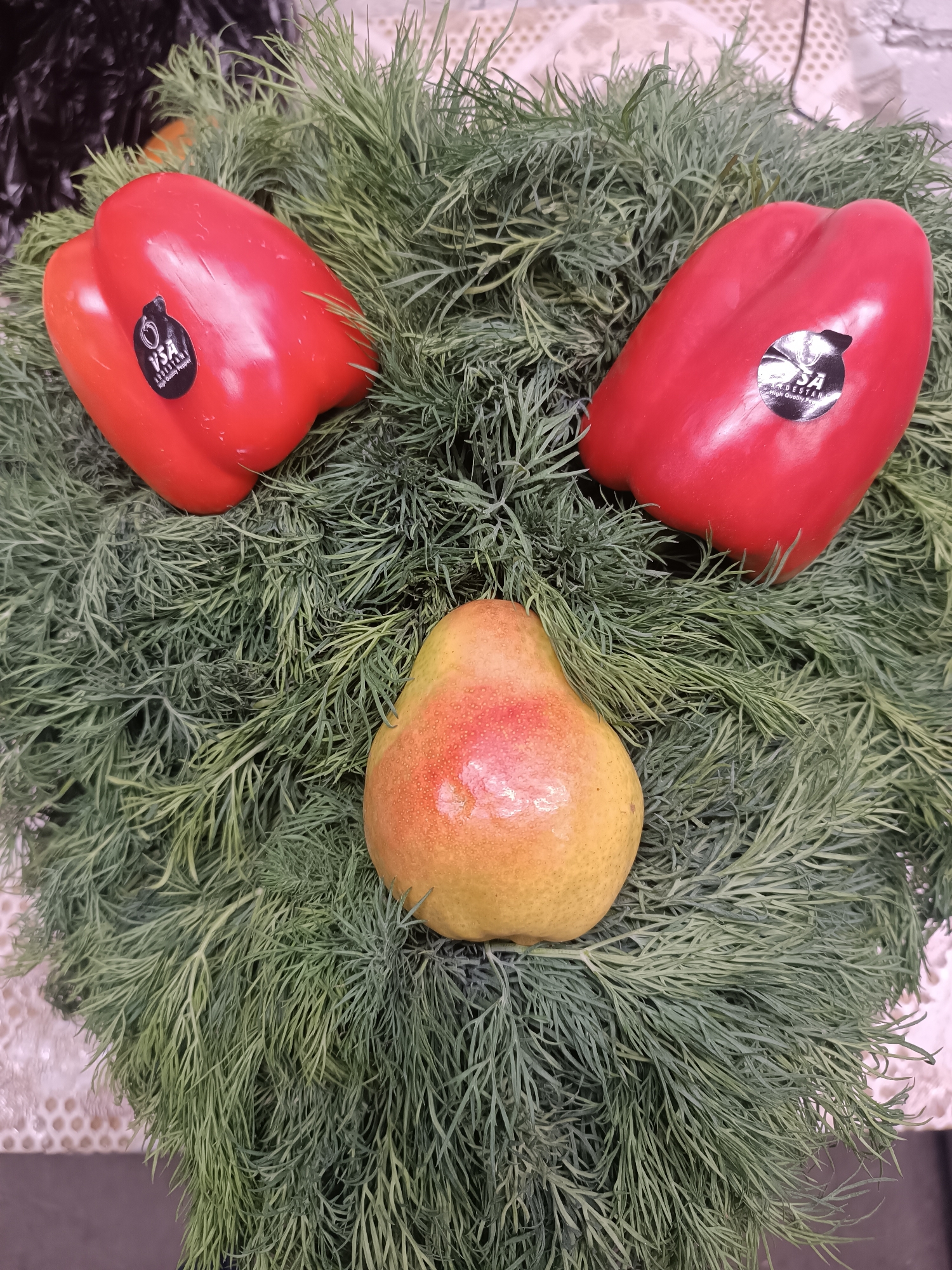 A still life composition features two red bell peppers and a pear arranged on a bed of dill. The peppers are positioned at the upper left and upper right, respectively, with stickers still affixed. The pear is centrally located beneath them. The location is indicated by the presence of stickers from a vendor "VSA Andesstan" with a possible Chaikovsky, Russia affiliation.