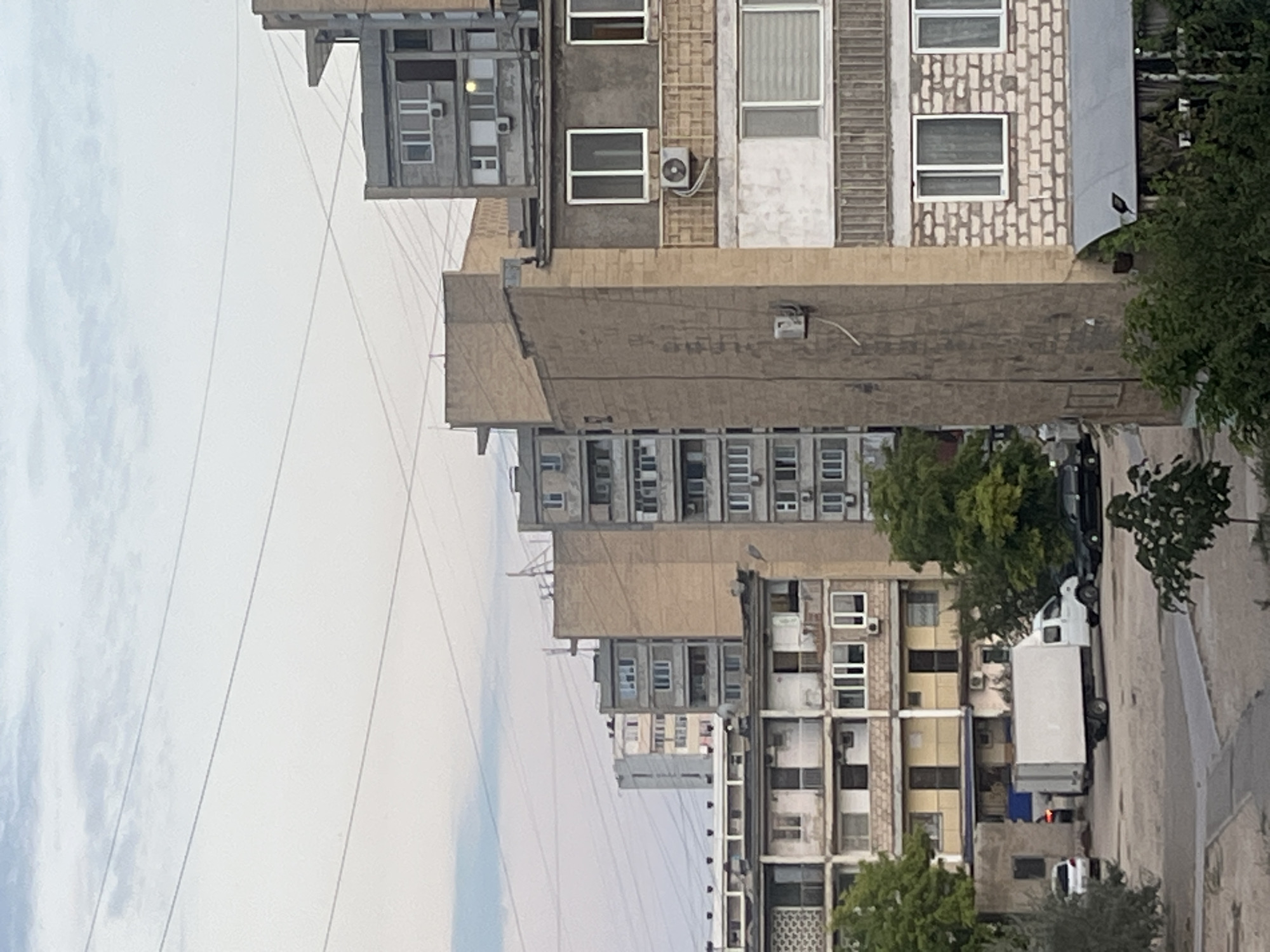 The image depicts a street scene in Aktau, Kazakhstan, characterized by a row of apartment buildings and a street with vehicles. The perspective is from a low angle, looking upwards towards the buildings, which are predominantly concrete structures with numerous windows. The buildings display a functional, perhaps Soviet-era, architectural style, with balconies and external air conditioning units visible.
In the foreground, a white truck with a large cargo box is parked on the right side of the street. To the left of the truck, a dark-colored car is partially visible. Further down the street, another white vehicle, possibly a smaller car, is parked. Trees with green foliage are interspersed along the street and between buildings, adding touches of nature to the urban environment.
The sky is overcast, suggesting a cloudy day, and the overall lighting indicates it is likely daytime. The scene appears calm and ordinary, with no specific event or activity prominently taking place. The focus is on the architecture and the streetscape. No legible text is discernible in the image. The orientation of the image is rotated 90 degrees counter-clockwise.