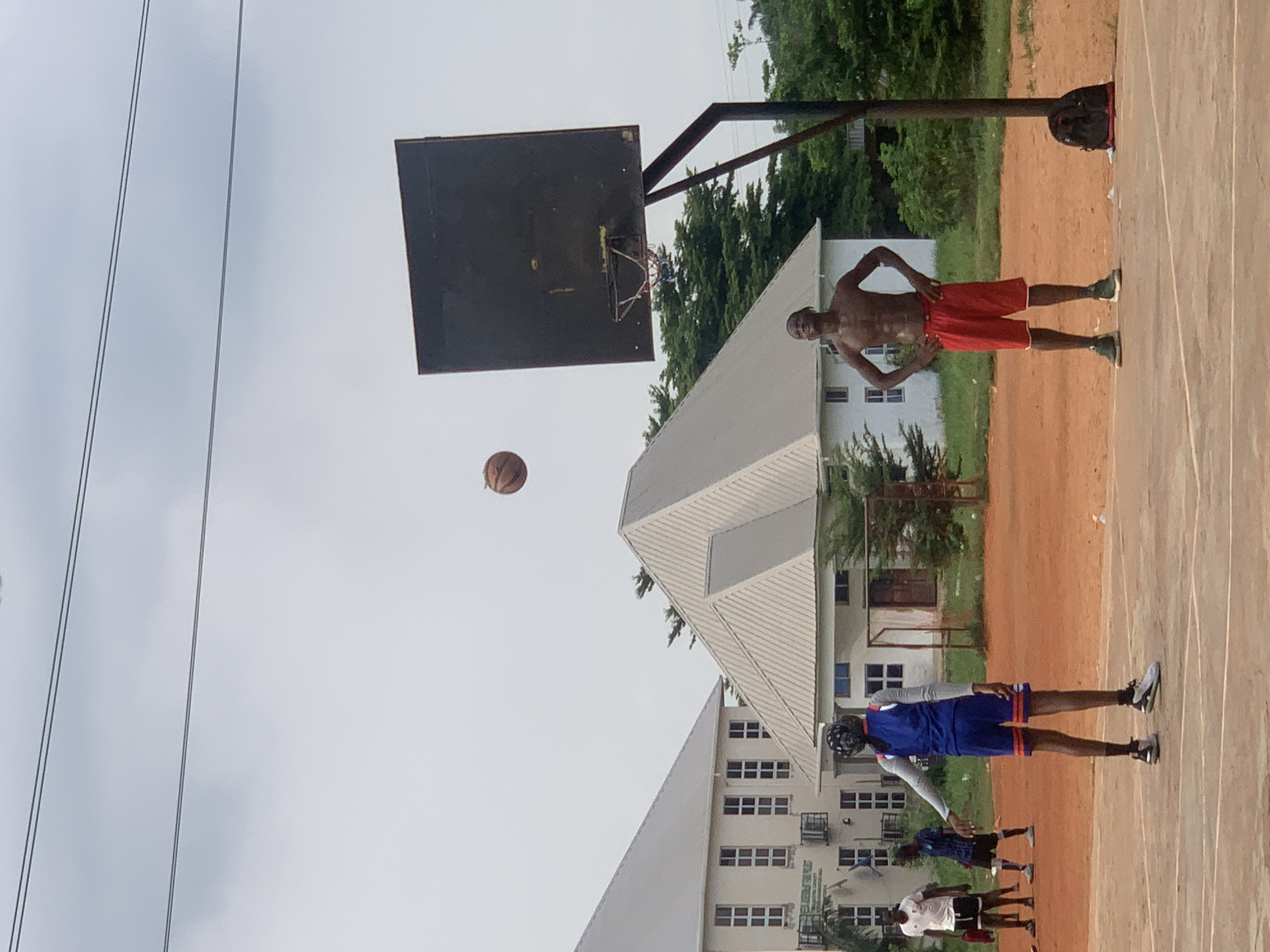 In Ukukwa, Nigeria, on an outdoor basketball court with a reddish-brown dirt surface, a group of young men are engaged in a game. The sky is overcast. The court is bordered by some trees and two large, white buildings with grey, gabled roofs. One of the buildings has multiple windows arranged in rows. The other building has a single, pitched roof. A basketball hoop with a black backboard and a red, white, and blue net stands on the right side of the frame. A black backpack rests on the ground near the base of the hoop.
In the foreground, two individuals are the primary subjects. On the left, a person wearing a blue and orange basketball uniform, complete with a bandana on their head, faces away from the camera, their left arm extended. To the right, a shirtless young man with muscular build stands with his hands on his hips, wearing red basketball shorts and green sneakers. A basketball is in mid-air above and to the left of the hoop, suggesting it has just been shot. In the background, on the left side of the court, two other young men are visible. One is wearing a white t-shirt and black shorts, and the other is in a blue t-shirt and dark shorts. They appear to be observing the game.