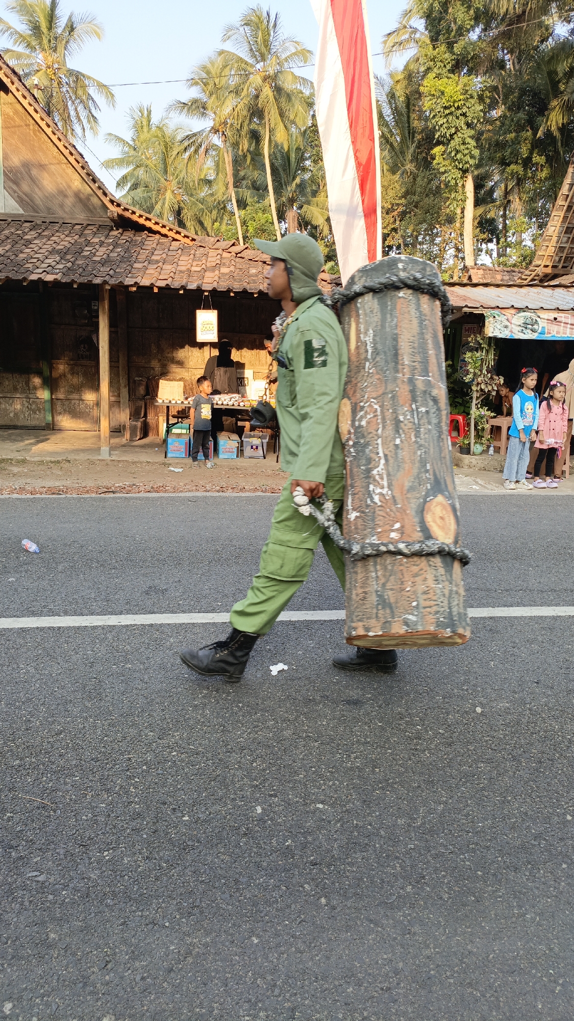 In Krajan Lor, Indonesia, on a paved road, a person in a green uniform and cap walks from left to right, carrying a large, cylindrical object resembling a tree trunk on their back. The object is secured with rope and chains. The person is wearing black boots and green cargo pants. Behind the person, a building with a tiled roof and open storefronts is visible, with palm trees and other vegetation in the background. A red and white Indonesian flag is displayed prominently. Several people, including children, are standing on the sidewalk near the storefronts. The scene appears to be a daytime outdoor event or procession.