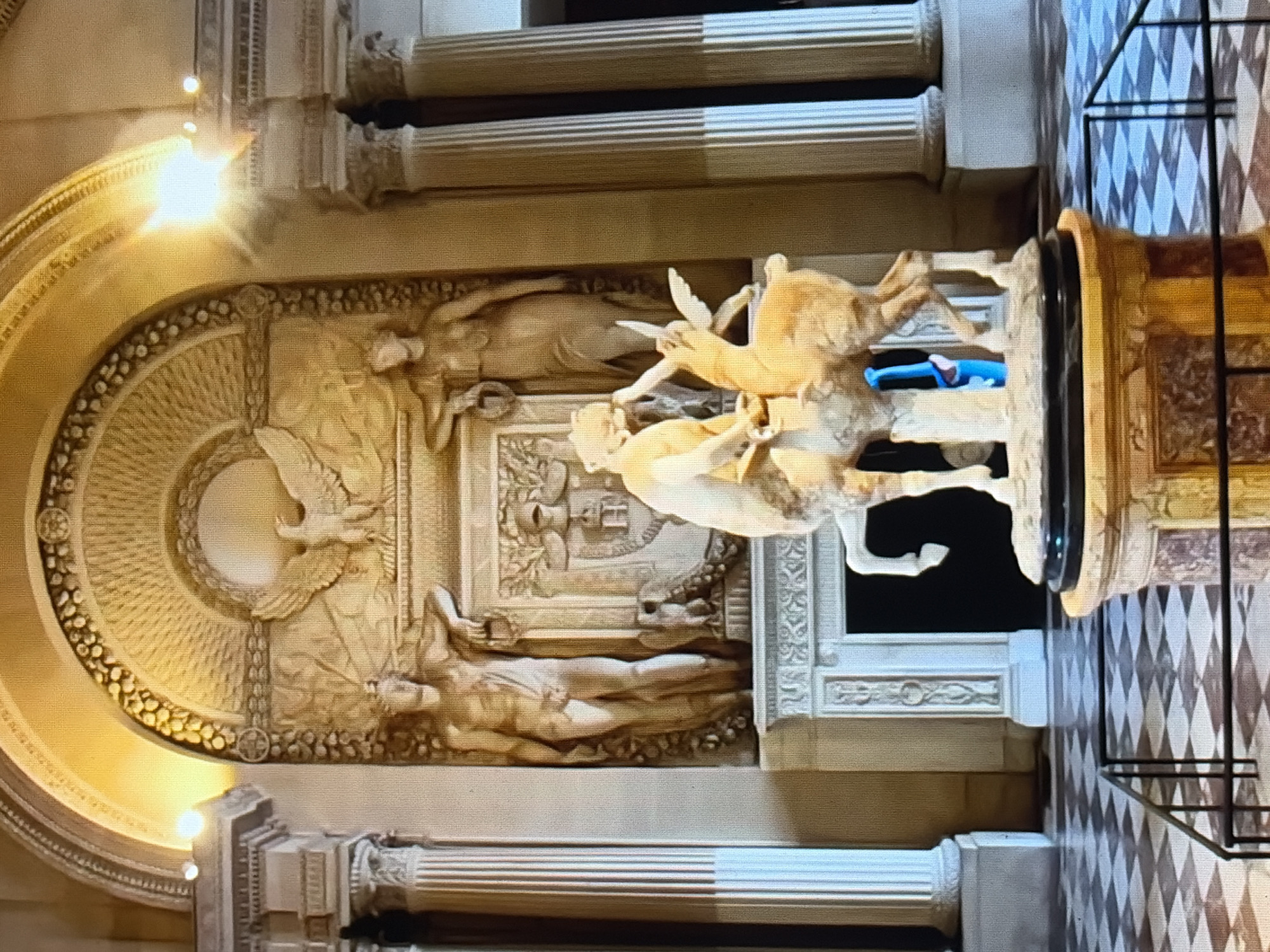 A marble sculpture of a satyr embracing a fawn is displayed in a grand hall. The satyr, a male creature with goat-like features, is depicted with human-like legs and a muscular torso, holding the fawn in a tender embrace. The fawn, a young deer, is shown with delicate wings, suggesting a mythical interpretation. Both figures are rendered in smooth, polished marble, with anatomical details meticulously carved.
The sculpture rests on a ornate pedestal with marble inlays, contributing to the opulent atmosphere of the hall. Behind the sculpture, a large, classical-style fireplace is set into a wall adorned with intricate relief carvings. Two imposing statues of male and female figures flank the fireplace, and above them, an eagle is depicted in mid-flight, surrounded by a laurel wreath. The hall features tall, fluted columns on either side, their grandeur emphasized by the soft, warm lighting that illuminates the space. The floor is a striking black and white checkered pattern, adding to the room's sophisticated aesthetic. A dark metal railing separates the viewer from the artwork, ensuring its preservation.