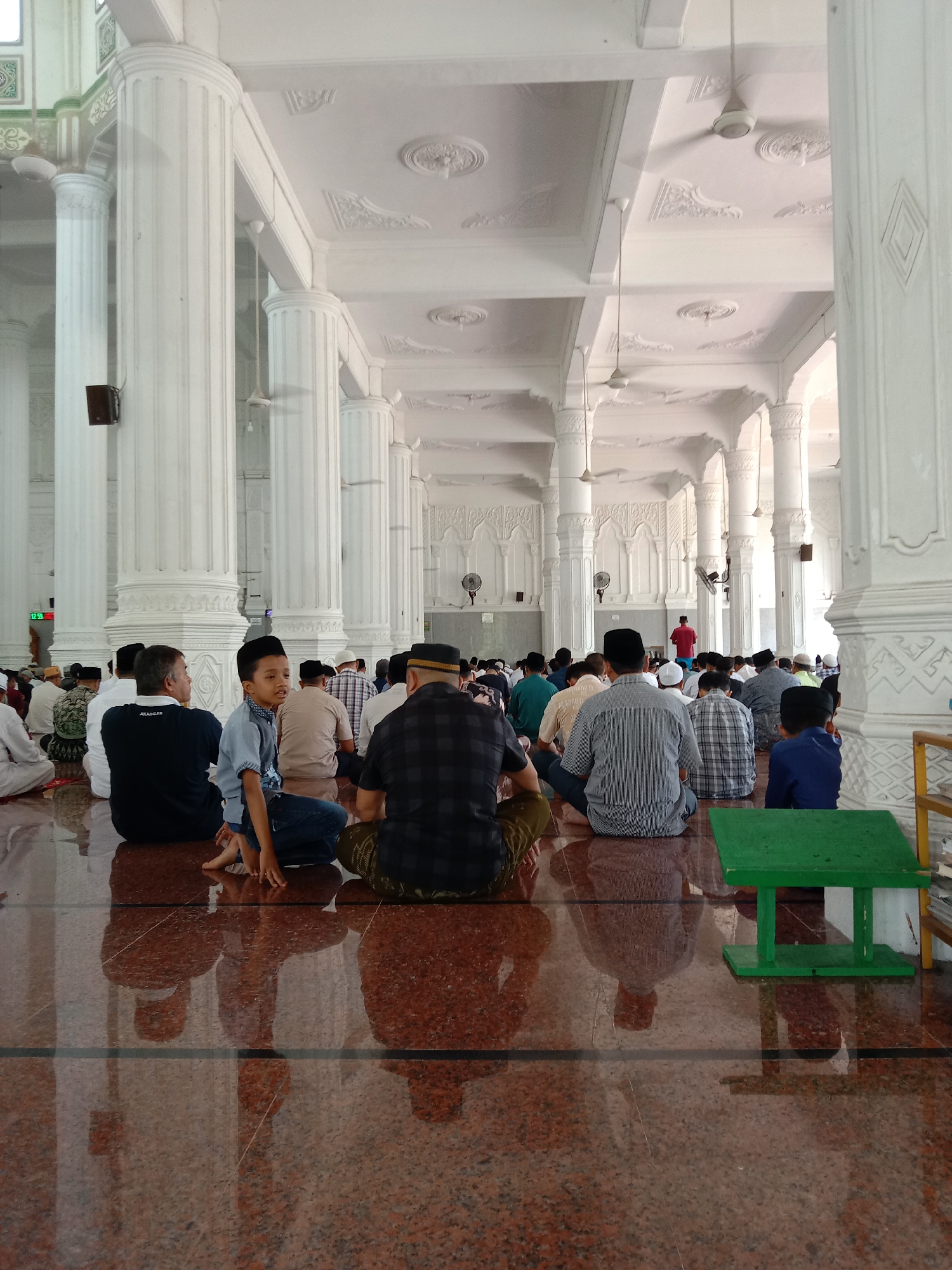 **Location:** A spacious prayer hall, likely inside Masjid Agung Islamic Center Lhokseumawe, Lhokseumawe, Indonesia.
**Environment:** The hall is characterized by predominantly white architecture, featuring numerous large, fluted columns supporting a high, ornately decorated ceiling. Ceiling fans are visible. The floor is polished with a brown, speckled pattern.
**People:** A group of Muslim men and boys sit on the floor, facing away from the camera, in preparation for prayer. They are dressed in a variety of casual and traditional attire, including shirts, pants, and some wearing skullcaps.
**Actions:** The individuals are seated and appear to be waiting for prayer to commence. Their posture suggests attentiveness and reverence.
**Objects:** A small green stool stands to the right of the frame.
**Interactions:** The people are coexisting peacefully and silently, awaiting the start of their communal prayer.