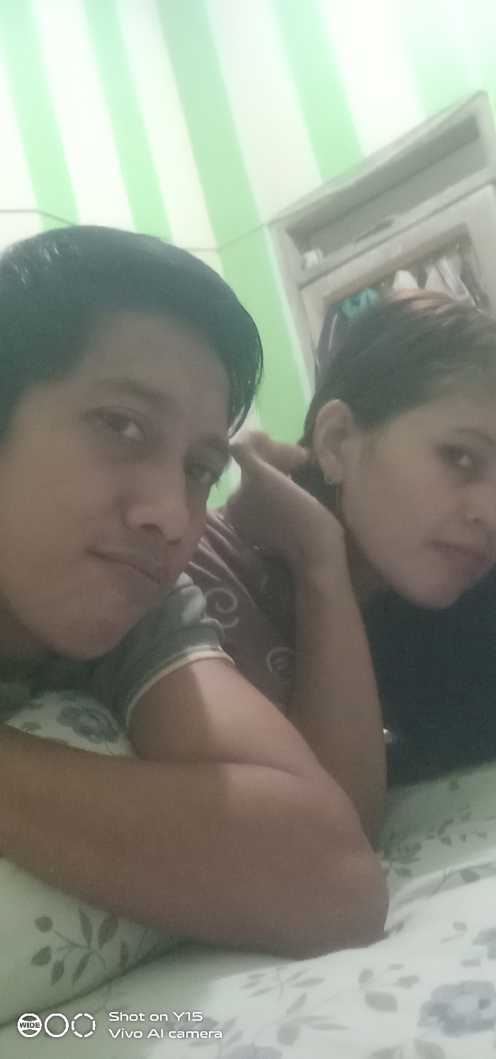The image displays two individuals positioned indoors in Ranugedang, Indonesia. The subject in the left foreground has dark, short hair and is looking directly at the camera. Their right arm is bent, supporting their head, and they are wearing a light-colored, possibly striped, short-sleeved top. A second individual is partially visible behind and to the right, possessing darker, longer hair and wearing a dark-colored top. This individual is also oriented towards the camera, with a small earring visible on their right ear. Both individuals are lying or reclining on a surface covered by a light-colored sheet with a subtle floral pattern. The background features a wall decorated with alternating vertical light green and white stripes. In the upper right quadrant, a portion of an open cabinet or shelving unit, characterized by a light frame and a darker interior, is visible. The lower-left corner of the image contains the metadata "Shot on Y15 Vivo AI camera."