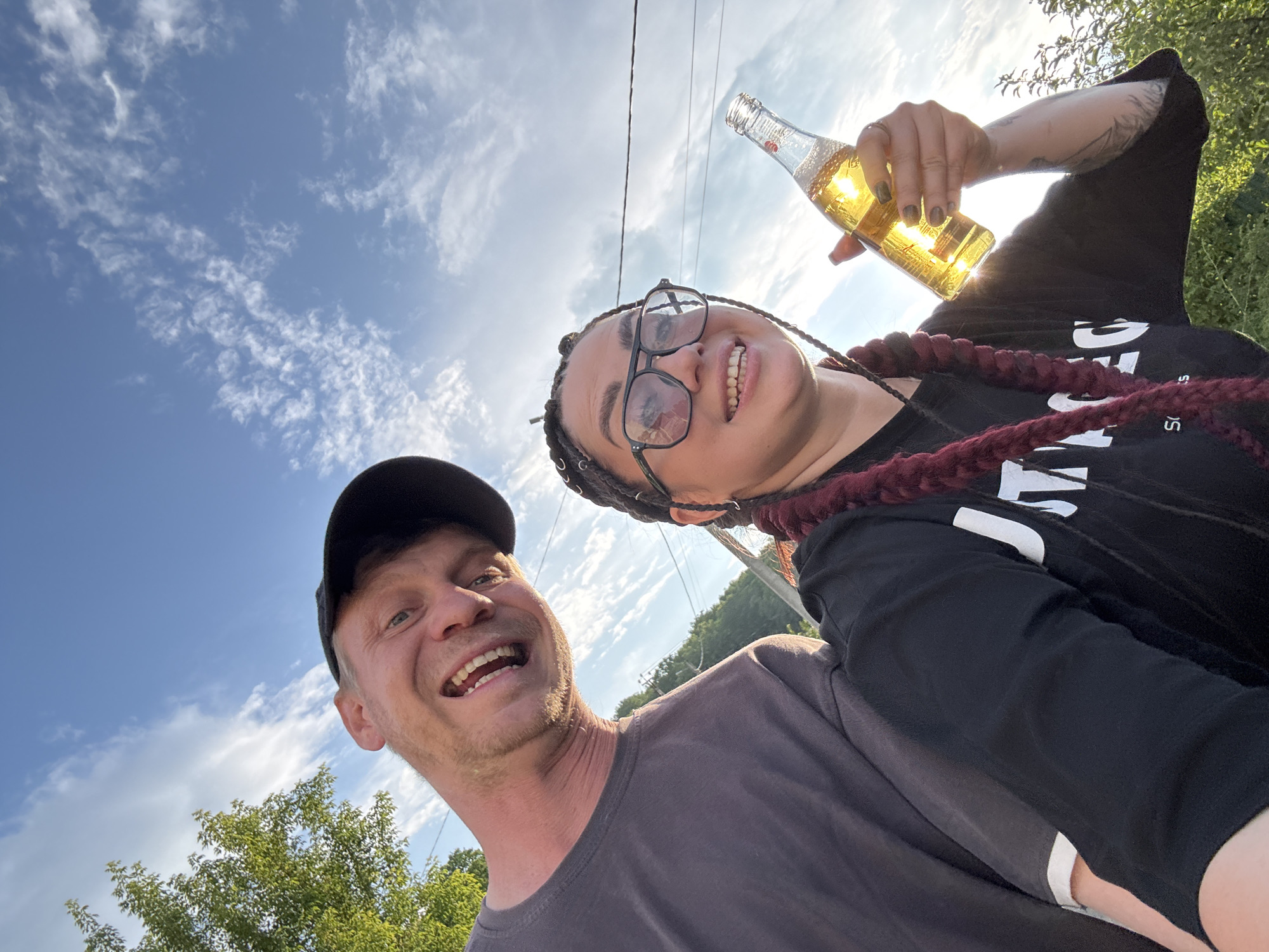 In this full-frame, outdoor, waist-up selfie, a Caucasian man and a woman are posed from a low angle against a bright blue sky with wispy clouds. The man, on the left, is wearing a dark grey t-shirt and a black baseball cap, with his head tilted and a wide smile. The woman, on the right, has dark braided hair, is wearing black-rimmed glasses and a black t-shirt. She is holding a clear glass bottle filled with a yellow liquid, which is illuminated by the sun. Her left hand is visible, with manicured nails. Tattoos are visible on her right arm. The background consists of greenery and a portion of a white building. The scene is set in an outdoor environment, possibly a rural or suburban area, under a bright, daytime sky.