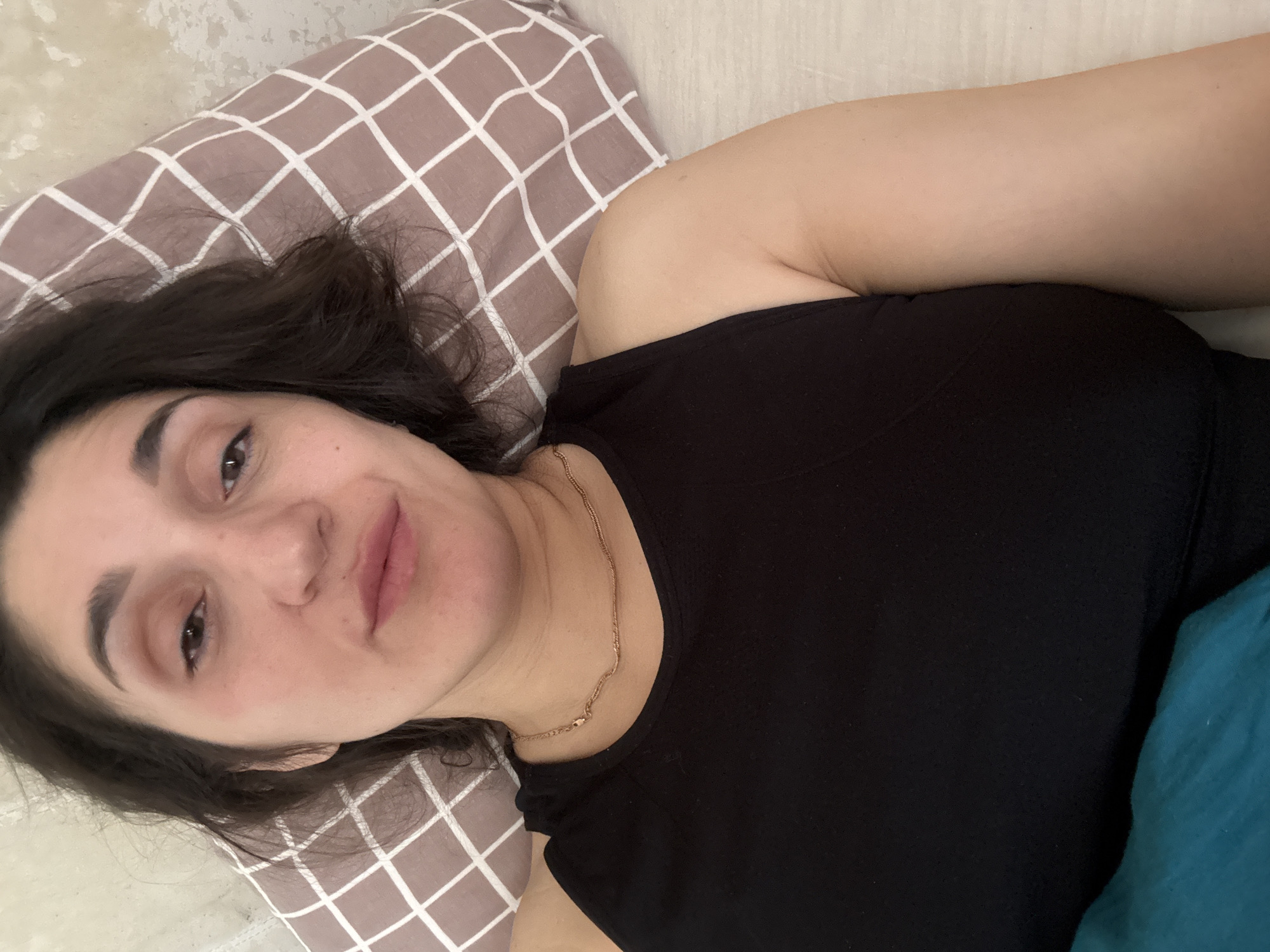 A woman with dark hair is lying on a bed. She is wearing a black tank top and a gold necklace. The bed has a pillow with a brown and white checkered pattern. The environment is indoors, and it is not possible to determine the location as Tobolsk, Russia from the image.