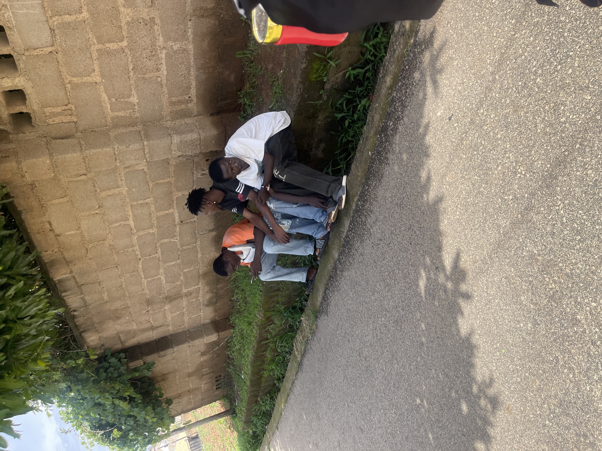 Three young men are seated on a low concrete curb along a roadside in Jos, Nigeria. The man on the left, wearing an orange t-shirt and light blue jeans, is looking at his phone. The middle individual, in a black t-shirt with white stripes, has his head turned to the left. The man on the right, dressed in a white t-shirt and dark pants, is looking down and to his right. Behind them is a rough brick wall, partially covered with green vines, and beyond that, more greenery. The road in front of them is paved asphalt, with shadows of leaves cast upon it. A red object with a yellow lid is visible on the right edge of the frame, held by an unseen person. The environment suggests a sunny day in an urban or semi-urban setting.