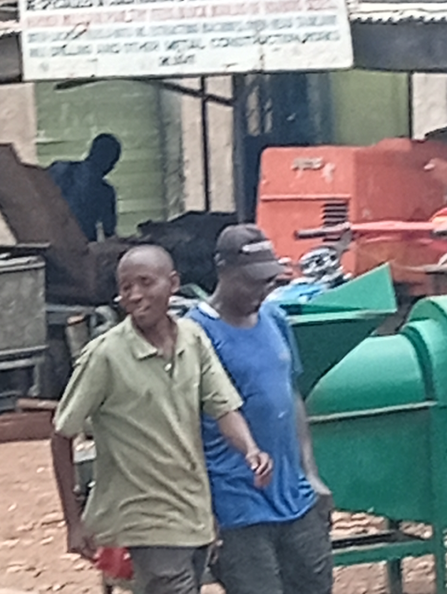 The image depicts an outdoor scene in what appears to be an industrial or workshop area in Gombe, Nigeria. In the foreground, two men are visible, walking or standing. The man on the left is wearing a light green collared shirt and dark pants. He has a shaved head and is looking towards the viewer with a slight smile. The man on the right is wearing a blue t-shirt and a dark baseball cap. He is facing to the left, and his expression is not clearly discernible.
Behind the men, various objects are present. To the left, a dark silhouette of a person is visible, potentially working or observing. Further back, there are large, industrial machines, including an orange structure that resembles a generator or compressor with metal grilles, and several large, green metallic objects that might be parts of machinery or storage containers. A sign with text in white is mounted above the scene, but the text is partially obscured and blurry, making it difficult to read clearly. The ground appears to be dirt or gravel. The overall impression is that of a busy commercial or manufacturing setting.