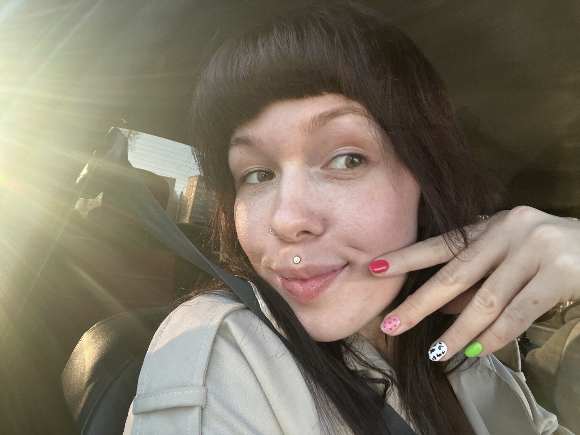 A woman is in a car, wearing a seatbelt. She has dark hair and bangs, a nose ring, and a lip piercing. Her nails are painted in different designs: red, pink with hearts, cow print, and green. The sun is shining brightly, visible through the car window, and an apartment building is seen in the background, outside the car. The location is Chaikovsky, Russia.