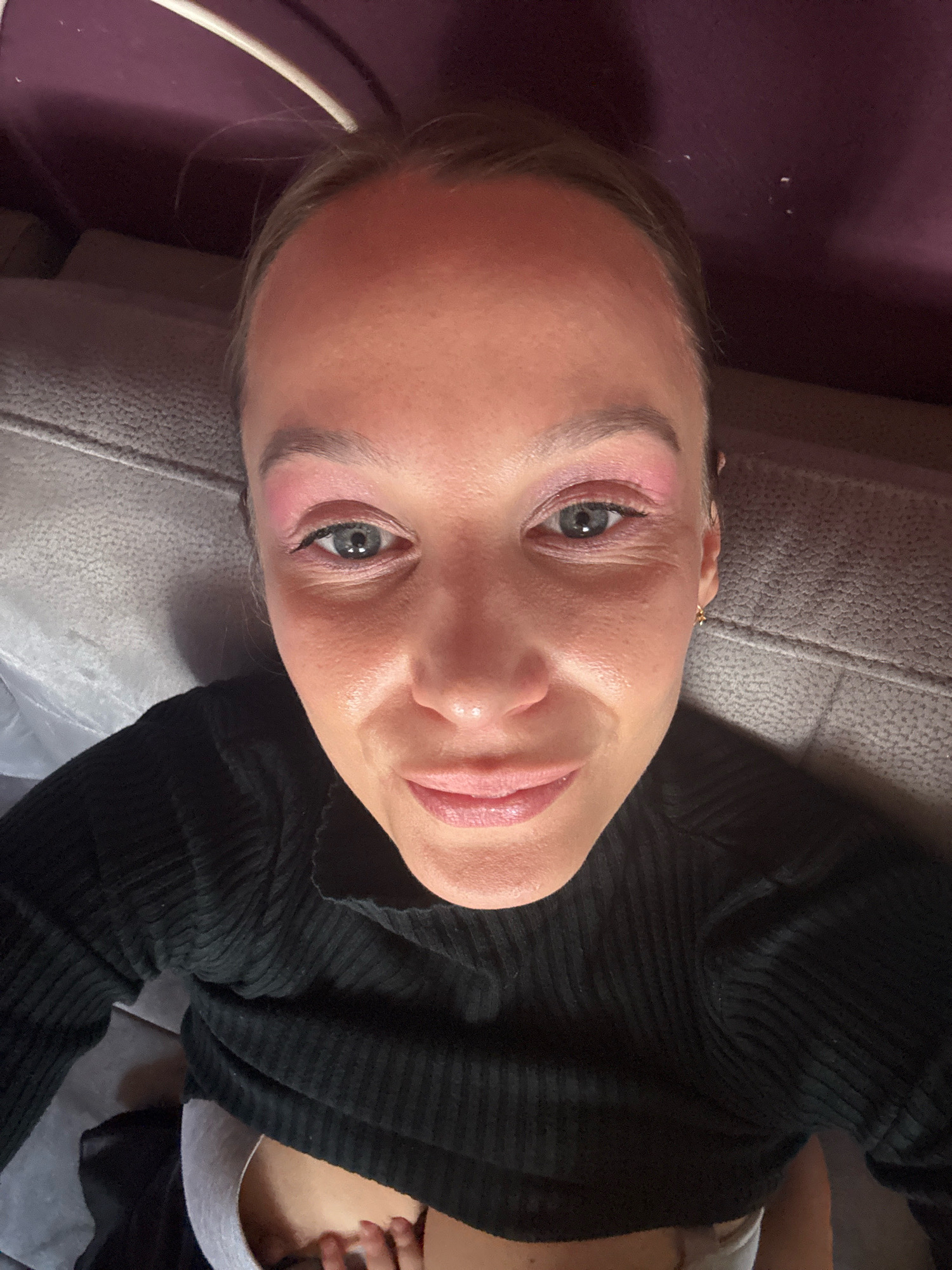 A close-up, selfie-style image shows a young woman lying down, looking up at the camera. She is wearing a black, ribbed, long-sleeved shirt and what appears to be light-colored underwear. She has pink eyeshadow on her eyelids and a light coating of makeup on her face. Her lips are slightly parted, revealing a hint of teeth. The background suggests an indoor setting with a textured grey surface, possibly a sofa, and a purple, possibly fabric-covered wall or headboard. There is a white cord or strap visible at the top of the image. The lighting is somewhat dim, casting shadows on her face and highlighting the texture of her skin. The image does not contain any discernible landmarks or features that would indicate a specific location within Überherrn, Germany.