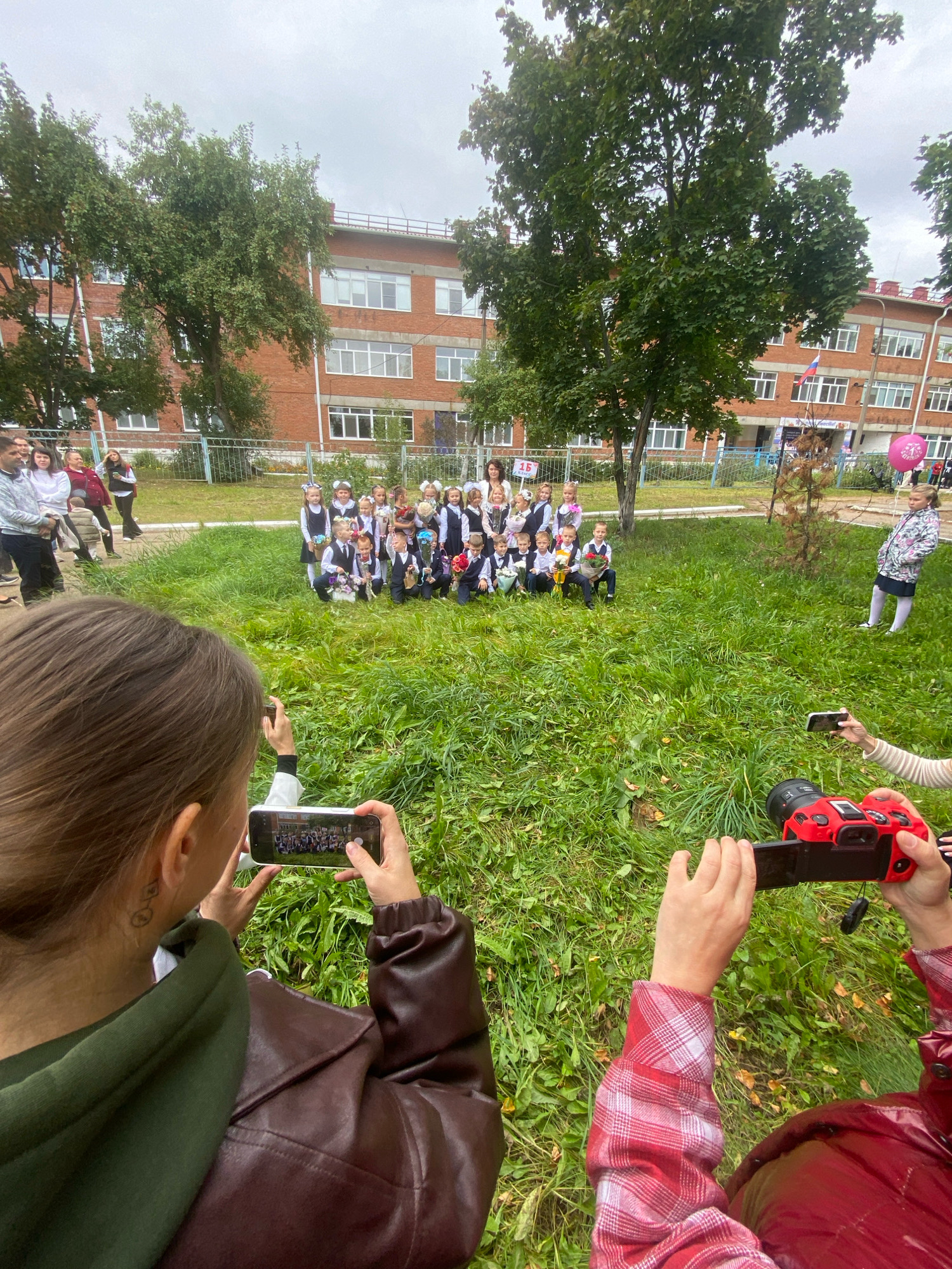 A group of children in school uniforms are posing for a photograph on a grassy area in front of a brick school building in Noviy, Russia. The children are holding bouquets of flowers. Two adults in the foreground are taking photos of the children with their phones and a camera. The sky is overcast.