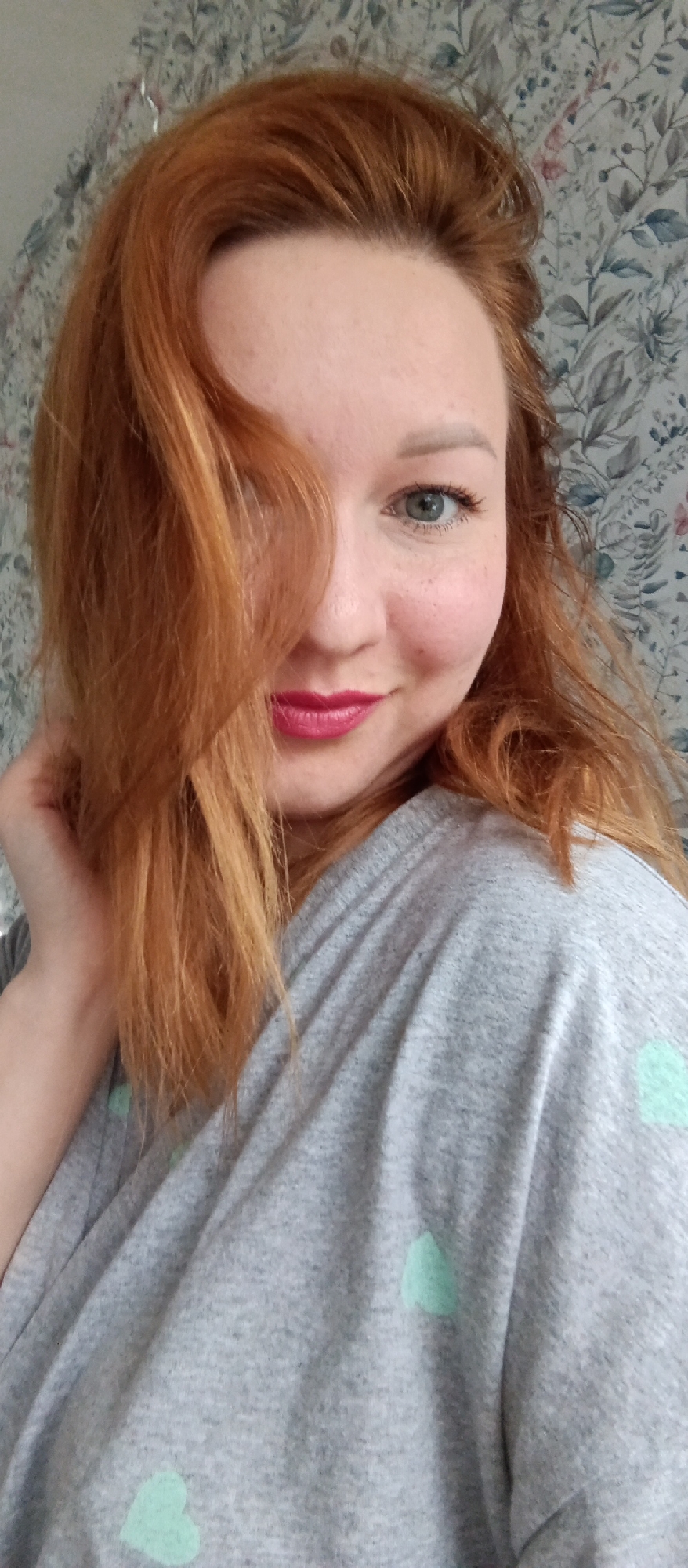 A selfie of a fair-skinned woman with red hair. The woman is wearing a grey shirt with light blue heart patterns. Her hair is partially obscuring her face. The woman is smiling slightly, with pink lipstick. The background is a wall with a floral pattern. The location is unknown.