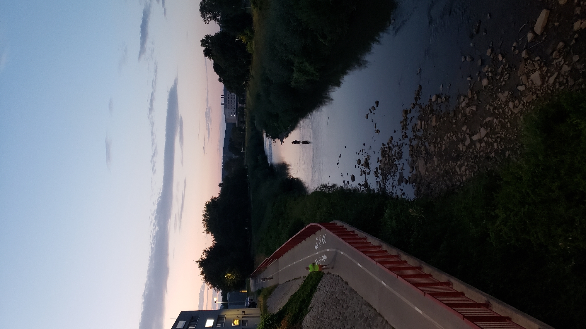 The image depicts a river flowing through a park-like area in Prešov, Slovakia, at dusk. A paved pathway with a red railing runs along the left bank of the river, bordered by a grassy embankment. On the pathway, two individuals are visible: one in a neon yellow shirt and another further back. In the river, a lone figure stands in the water, facing away from the camera, seemingly fishing or wading. The riverbed is rocky in places, with shallow water. The surrounding area is lush with green foliage and trees, with buildings visible in the background. The sky above is a gradient of pale blue to pink, indicating the setting sun.