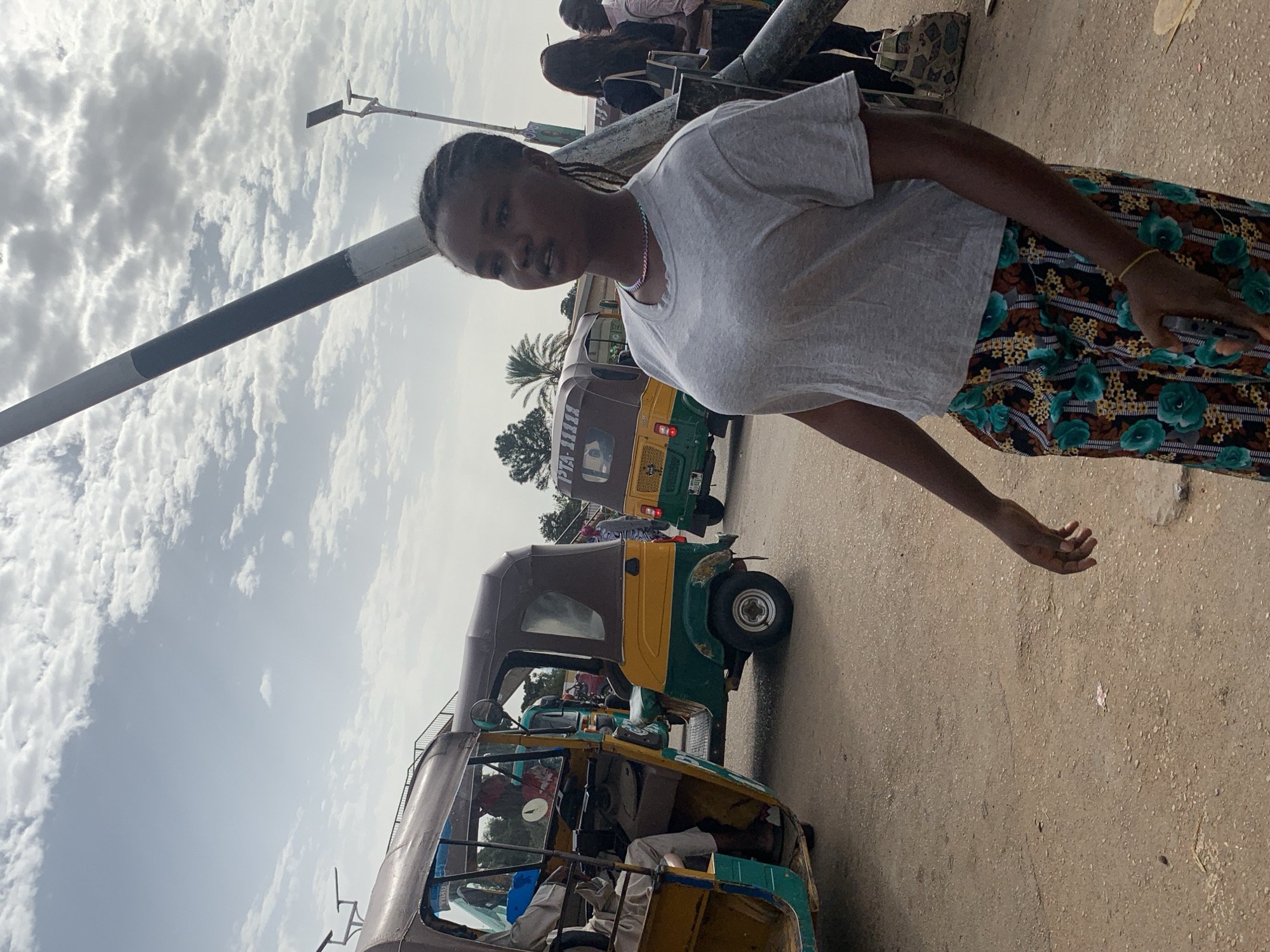 In Gura Top, Nigeria, a young woman with braided hair stands on a dirt road, wearing a grey t-shirt and a floral-patterned skirt. She is positioned in the foreground, slightly to the right of center, with her head turned to her left. Behind her, several auto-rickshaws, common in Nigerian cities, are visible. One auto-rickshaw, predominantly yellow and green, is parked to her left, with its door open. Another, a similar model, is further back and to the right, also in motion or parked. The background is a mix of sparse trees, a utility pole with a black and white striped band, and a cloudy sky. The scene appears to be a typical street or transportation hub.