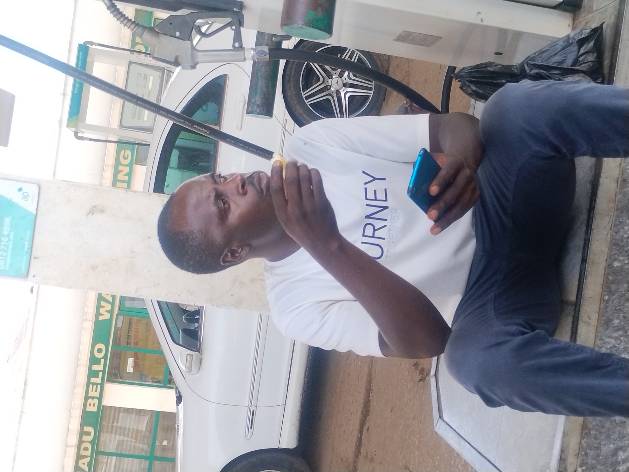 A man, appearing to be of African descent, is seated at a gas station. He is wearing a white t-shirt with "JOURNEY TOGETHER" printed on it and dark blue trousers. He is holding a blue smartphone in his right hand and appears to be looking at it. His left hand is raised towards his face, and he might be eating something.
The setting is a gas station, evident from the visible gas pump nozzle and hose. In the background, a white car, possibly a Mercedes-Benz, is parked, with its distinctive wheel visible. Another white car is partially visible on the left side of the image. The area is outdoors, and the lighting suggests it is daytime, though the specific time of day is not clear.
There is visible text on signs in the background. On the left, a sign with "ap" and a phone number "0812 716 4998" is present. Further to the right, a green sign reads "ADU BELLO WA" and "ING". Another sign on the pump reads "Polyhose FUEL", and "Energy for new world".
The overall mood seems relaxed, with the man engrossed in his phone. The scene depicts a common everyday activity at a service station.