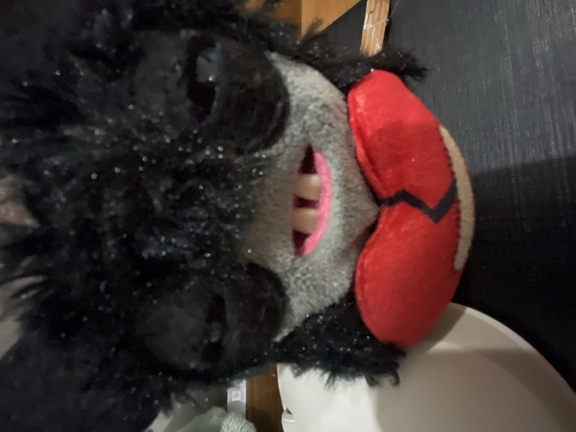 A close-up shot depicts a furry, plush toy head mounted on a white, possibly plastic, object. The toy head features a black, furry exterior with a gray face displaying teeth. A red, heart-shaped object with a black design is attached to the side of the head. The background includes a dark, textured wall and a wooden surface. The location in Denpasar, Indonesia is not deducible from the image.