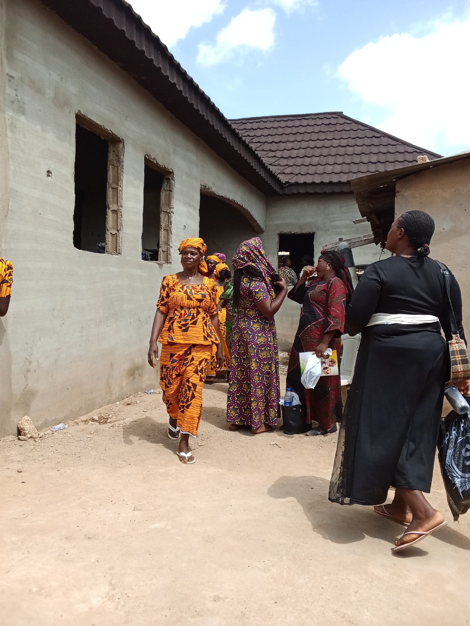 In Sabon Tasha, Nigeria, a group of women are gathered outdoors near a partially constructed building. The building has rough, light-colored walls and several unfinished window openings. A dark brown tiled roof is visible on the building. The ground is covered in dirt and sand, with some scattered debris.
The women are dressed in brightly colored, patterned traditional clothing, including headscarves. One woman in the foreground is wearing a vibrant orange and yellow patterned outfit with a matching headscarf, and she is walking towards the viewer. She has sandals on her feet. Other women in the background are also dressed in colorful attire, some with intricate patterns.
One woman in the background is wearing a dark red and black patterned dress and holding a white bag. Another woman, further in the background, is wearing a long, dark dress and a white sash. She is facing away from the camera and appears to be looking at something to her right. She is carrying a patterned bag over her shoulder and a plastic water bottle in her hand.
The sky above is partly cloudy, suggesting it is daytime. The overall scene appears to be a casual gathering or a moment between activities, possibly related to a local event or daily life. The expressions on the faces of the visible women suggest they are engaged in conversation or observation. There is no visible text in the image.