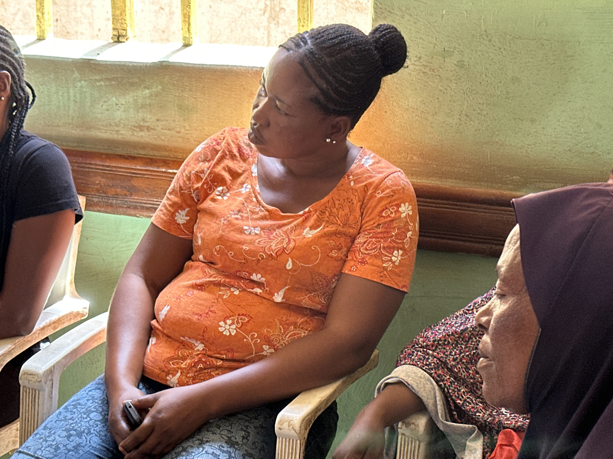 In an indoor setting, a woman with braided hair pulled back in a bun is seated on a white chair wearing an orange shirt with floral patterns and blue patterned pants. She holds a black object in her hands and looks to her left. To her left, a partial view of another person with braided hair is visible, while to her right, a person wearing a hijab and patterned garment can be seen. The background features a light green wall with a wooden molding and a window with light shining through. The location is Unknown, Unknown.