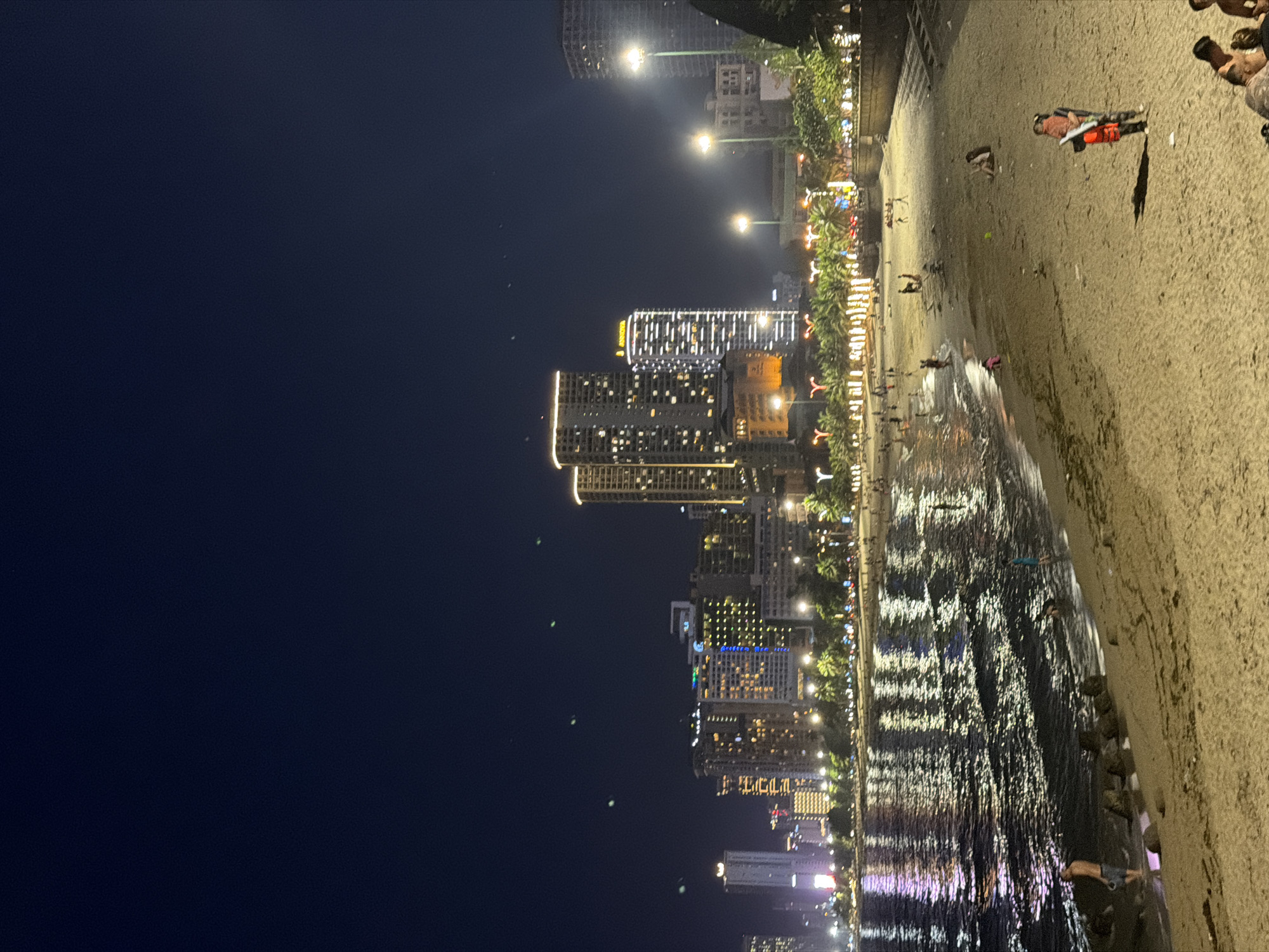 The image shows a nighttime view of a beach in Nha Trang Ward, Vietnam, with a cityscape in the background. The beach is sandy, and there are several people present, some walking, some standing in the shallow water, and some sitting on the sand. The ocean waves are reflecting the lights from the city, creating a shimmering effect. Palm trees are visible along the coastline, illuminated by decorative lights. Tall buildings, some with their windows lit, dominate the skyline. The sky is dark blue, suggesting it is night. In the foreground, a person is holding a child, and another person stands nearby. A life jacket is visible. There are also some scattered rocks in the water near the shore.