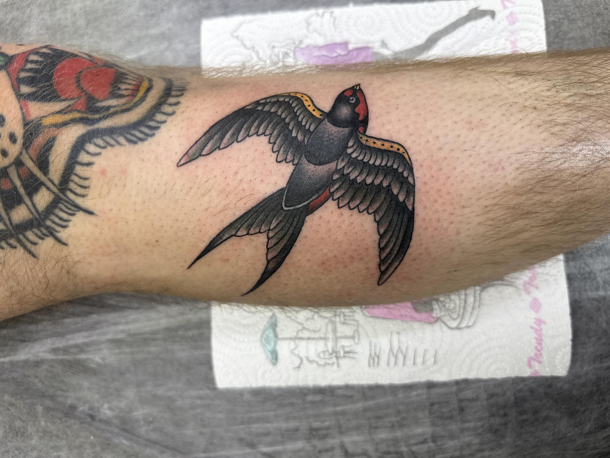 A forearm bears two tattoos: a traditional swallow in black, red, and yellow and a portion of another tattoo with red and yellow elements. The arm rests on a paper towel that reads "trendy tattoo" on a grey surface.