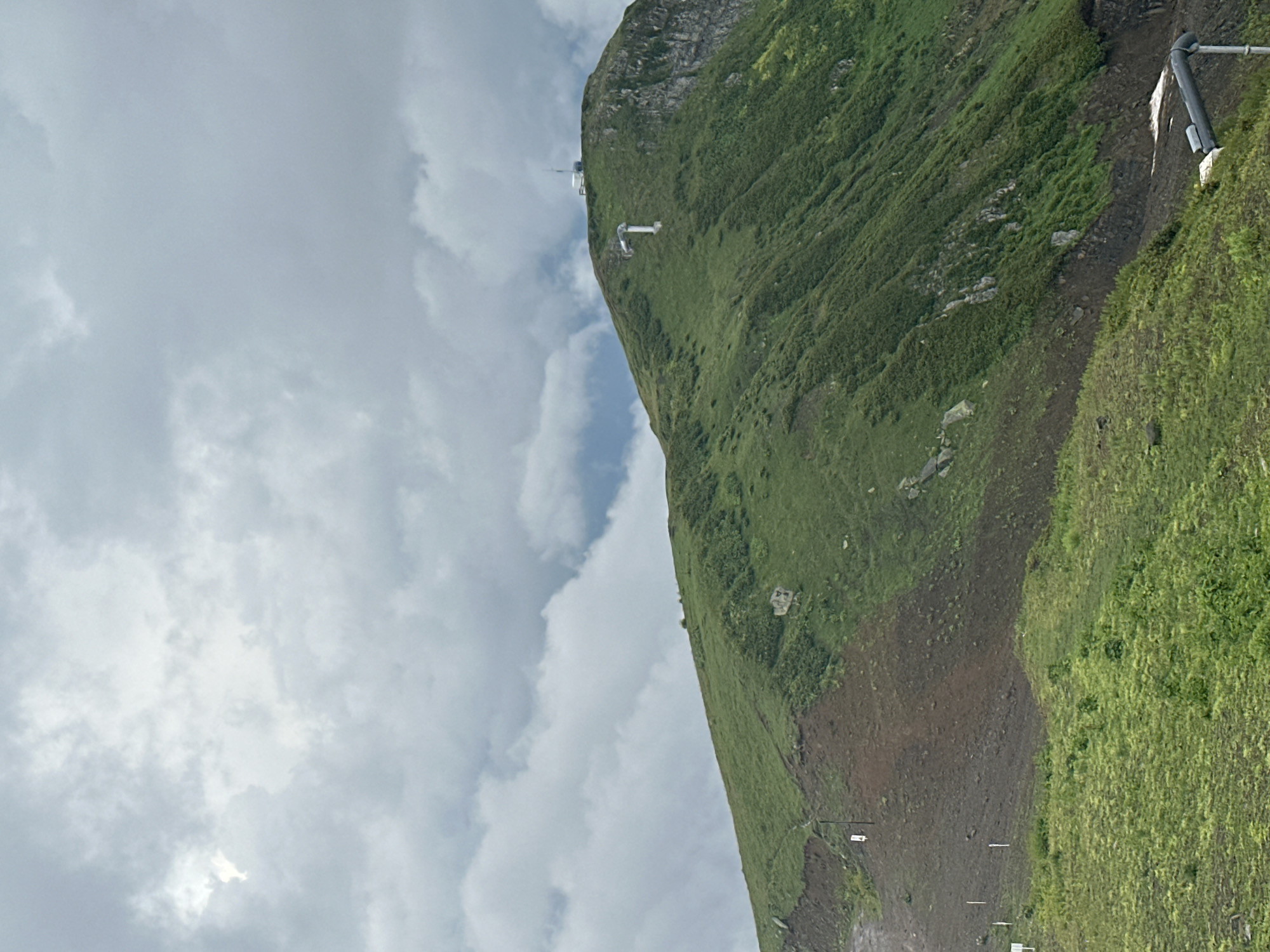 The image depicts a mountainous landscape under a cloudy sky in Estosadok, Russia. A verdant, grassy slope dominates the foreground and midground, with a steep ascent towards the summit. On the upper portion of the mountain, a ski lift structure is visible, with a tower and what appears to be a cabin or station at the peak. The terrain is characterized by lush green vegetation, with patches of exposed soil and rocks indicating erosion or different geological formations. The sky is overcast with heavy, grey clouds, suggesting a possibly cool or damp atmosphere. There are no people or animals present in the frame. The overall scene is one of natural beauty, with man-made infrastructure integrated into the mountain environment.